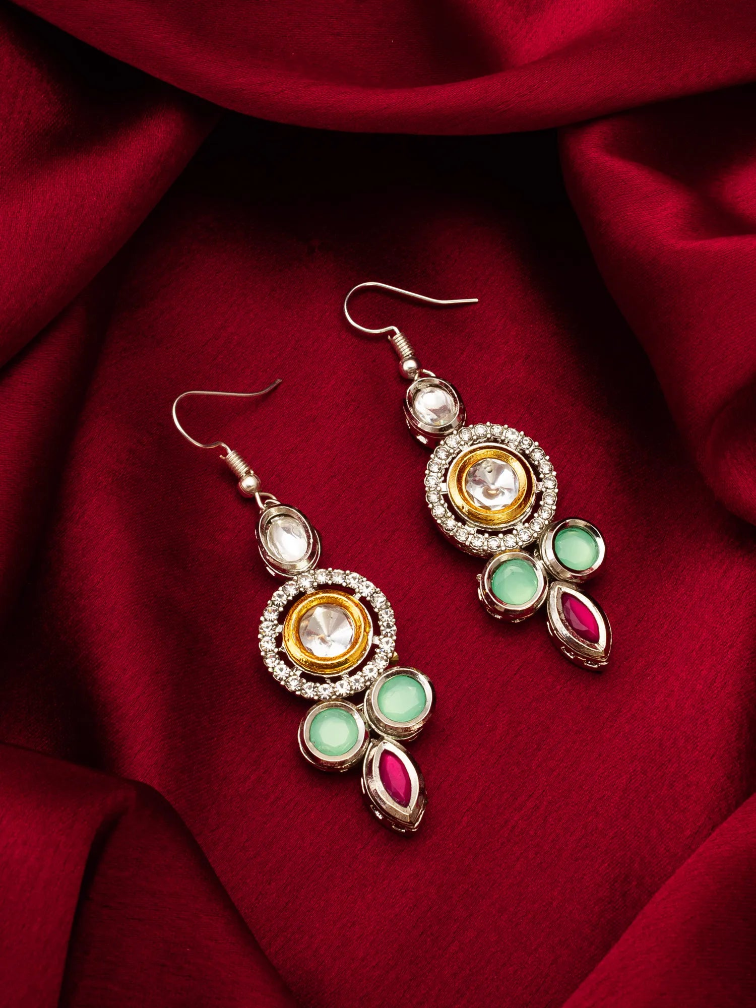 A pair of gold and silver dangle earrings with green and pink stones, displayed on a draped deep red fabric background.