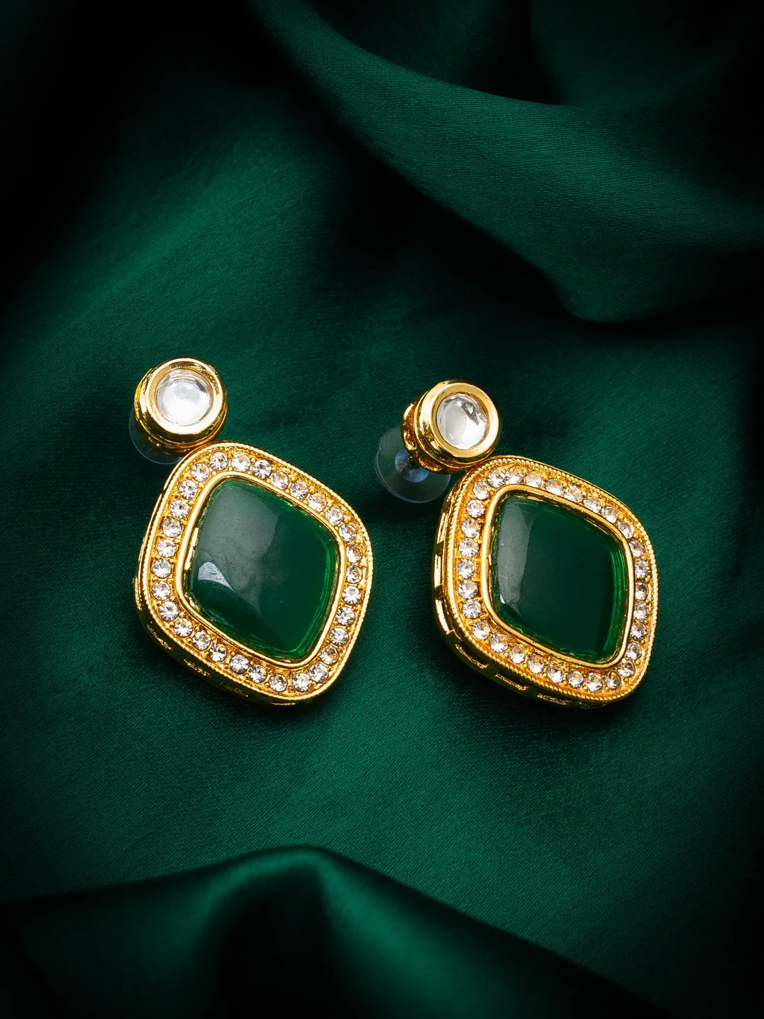 A pair of gold-plated statement earrings featuring large green marquise-shaped stones surrounded by small clear crystals, displayed on a dark green satin fabric background.