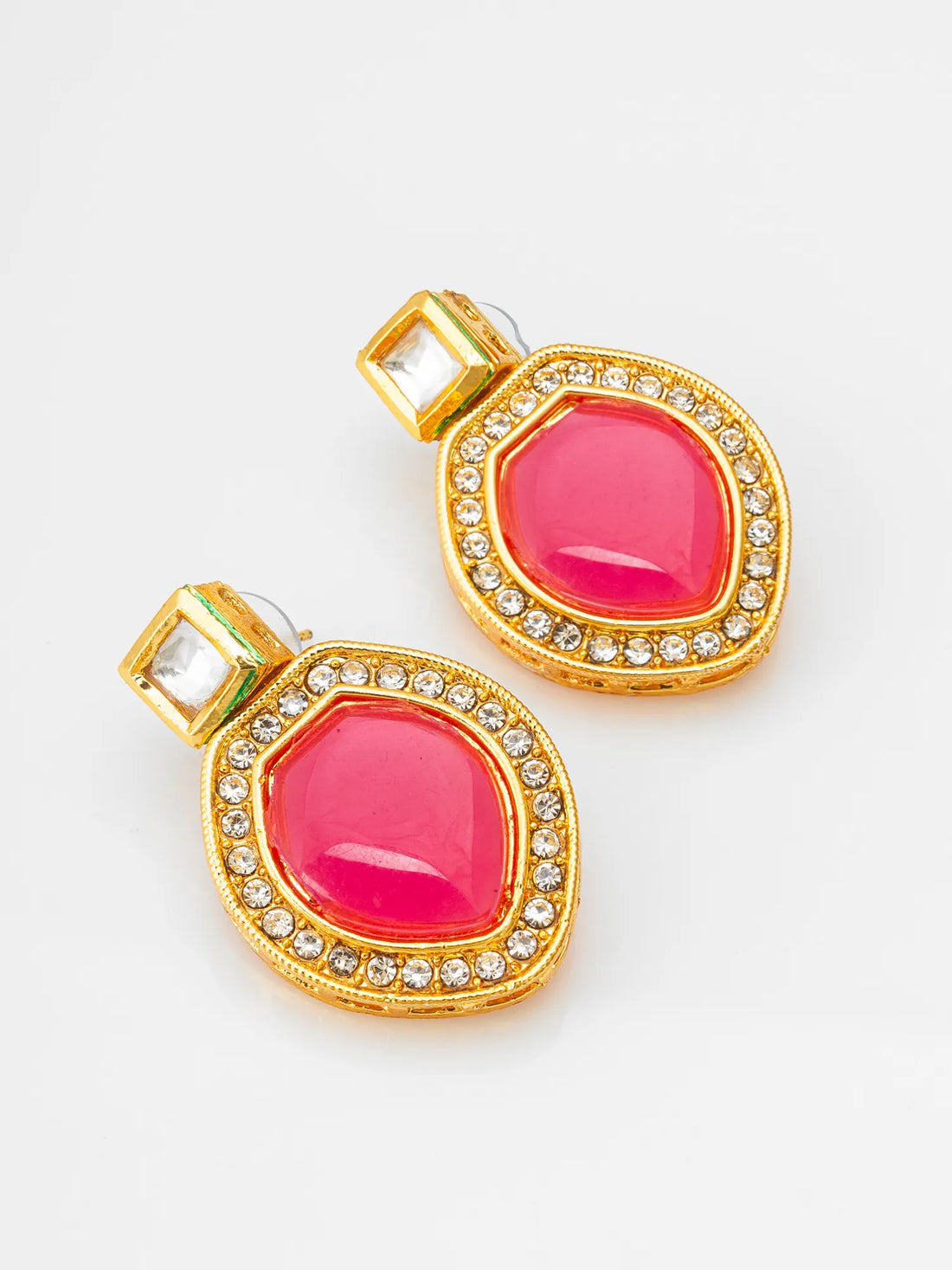 A pair of gold-toned statement earrings with large oval deep pinkish-red center stones and rows of small clear stones around the edges, displayed on dark green draped fabric.