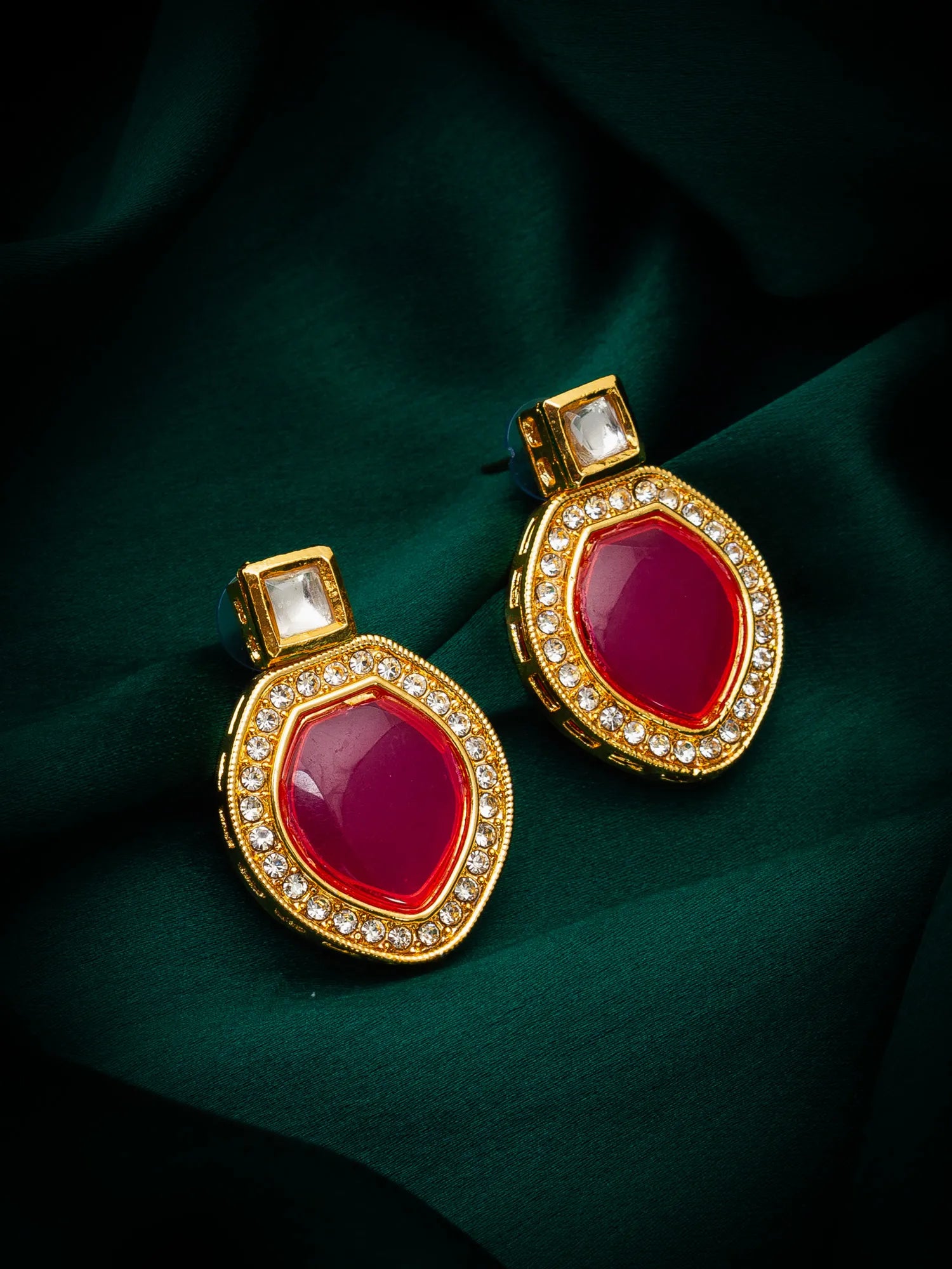 A pair of gold-toned statement earrings with large oval deep pinkish-red center stones and rows of small clear stones around the edges, displayed on dark green draped fabric.