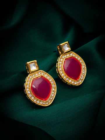 A pair of gold-toned statement earrings with large oval deep pinkish-red center stones and rows of small clear stones around the edges, displayed on dark green draped fabric.
