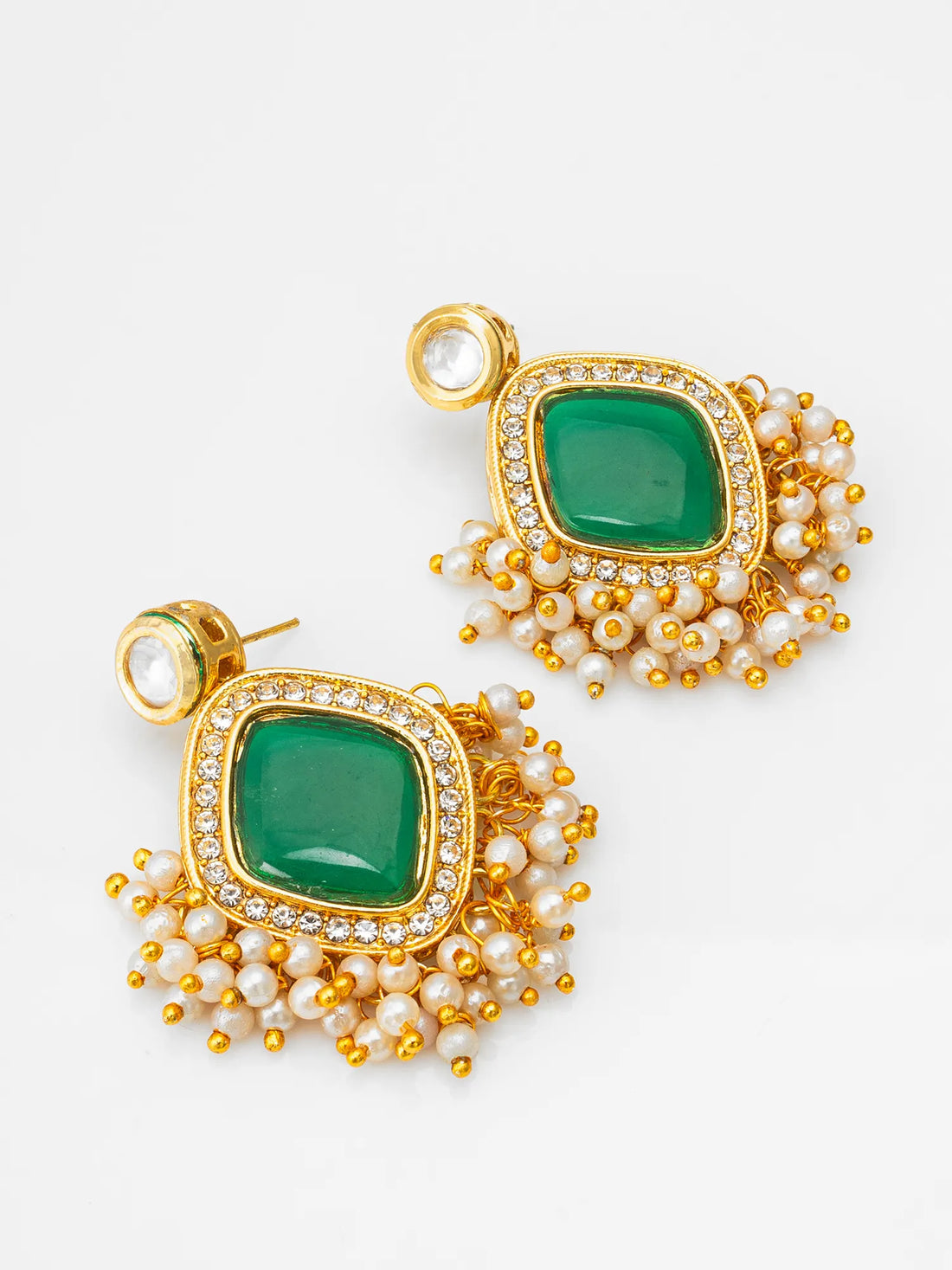 A pair of ornate gold-toned earrings with square green stone centers, surrounded by small clear stones and dangling pearl-like beads, displayed on dark green fabric.