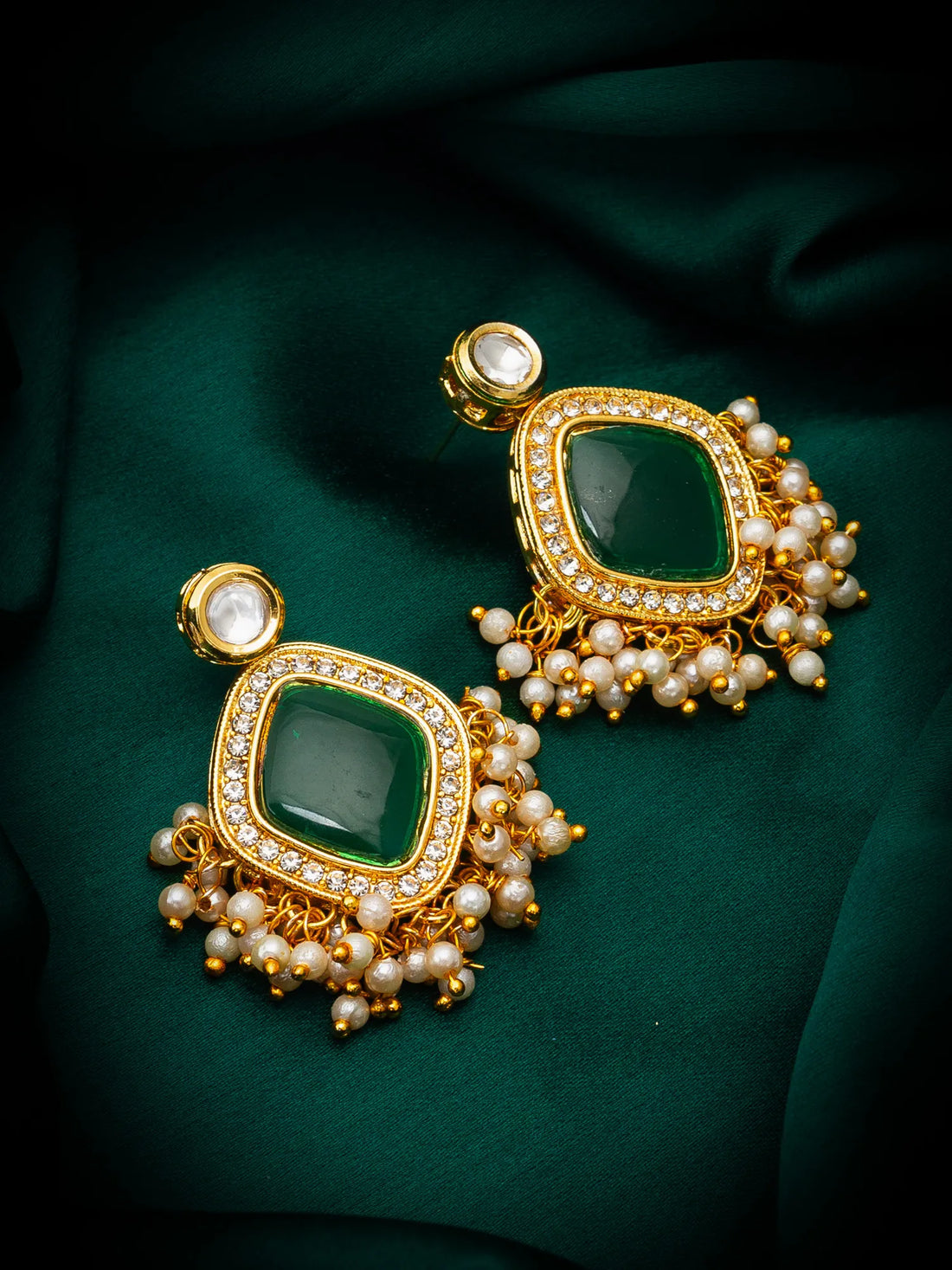 A pair of ornate gold-toned earrings with square green stone centers, surrounded by small clear stones and dangling pearl-like beads, displayed on dark green fabric.