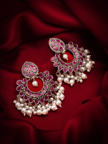 A pair of ornate silver-toned chandelier earrings embellished with pink stone stones and white pearls, displayed on a draped dark red satin fabric background.