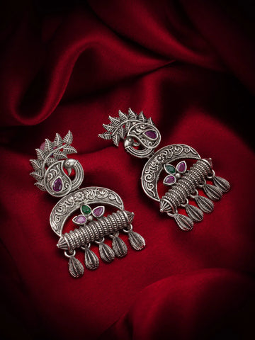 A pair of ornate silver-toned Indian jhumka earrings with paisley and floral motifs, accented with small red and green stones, displayed on a draped dark red satin fabric background.