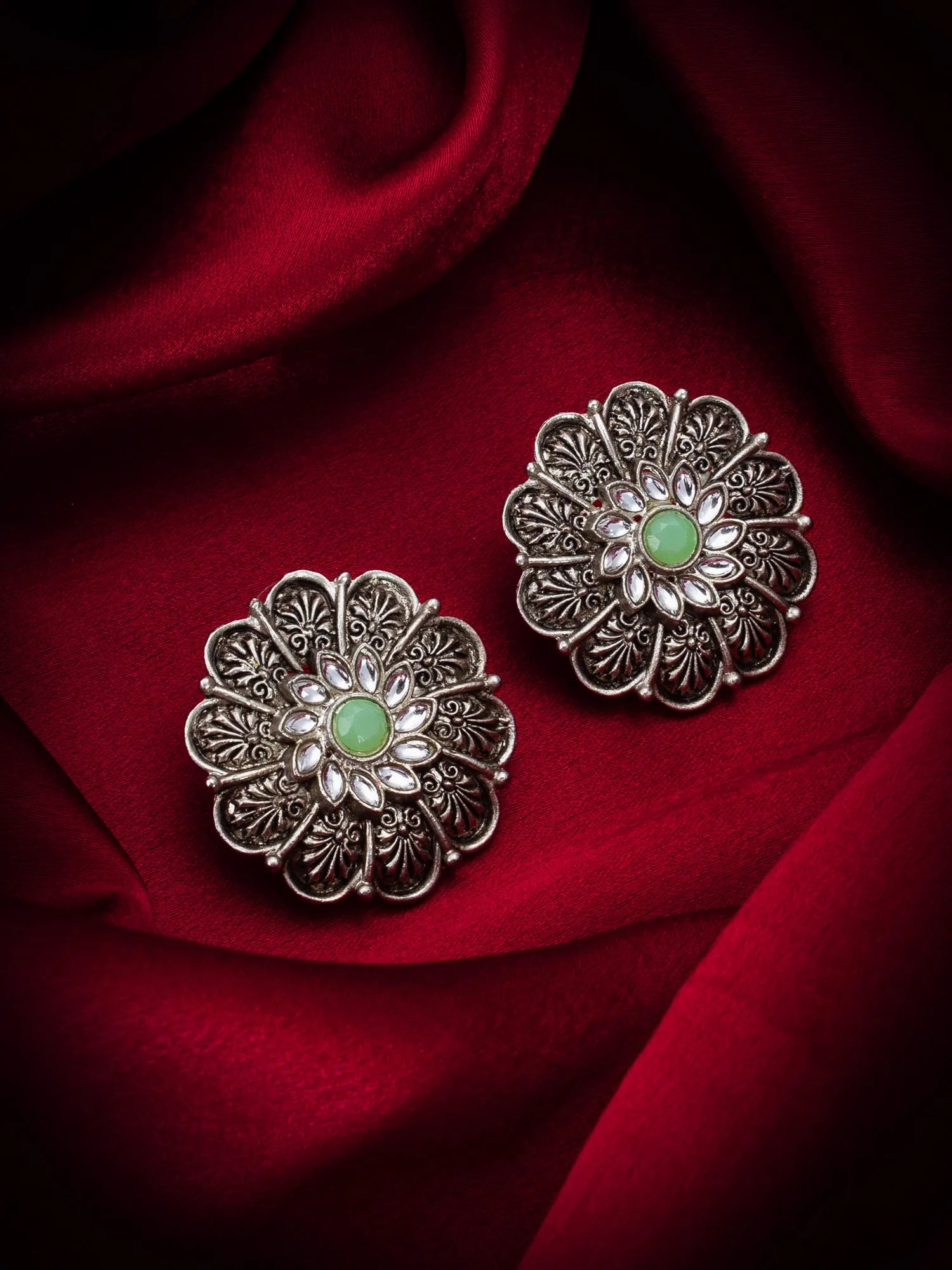A pair of silver-toned filigree flower-shaped earrings with oval green centers, displayed on a draped deep red fabric background.