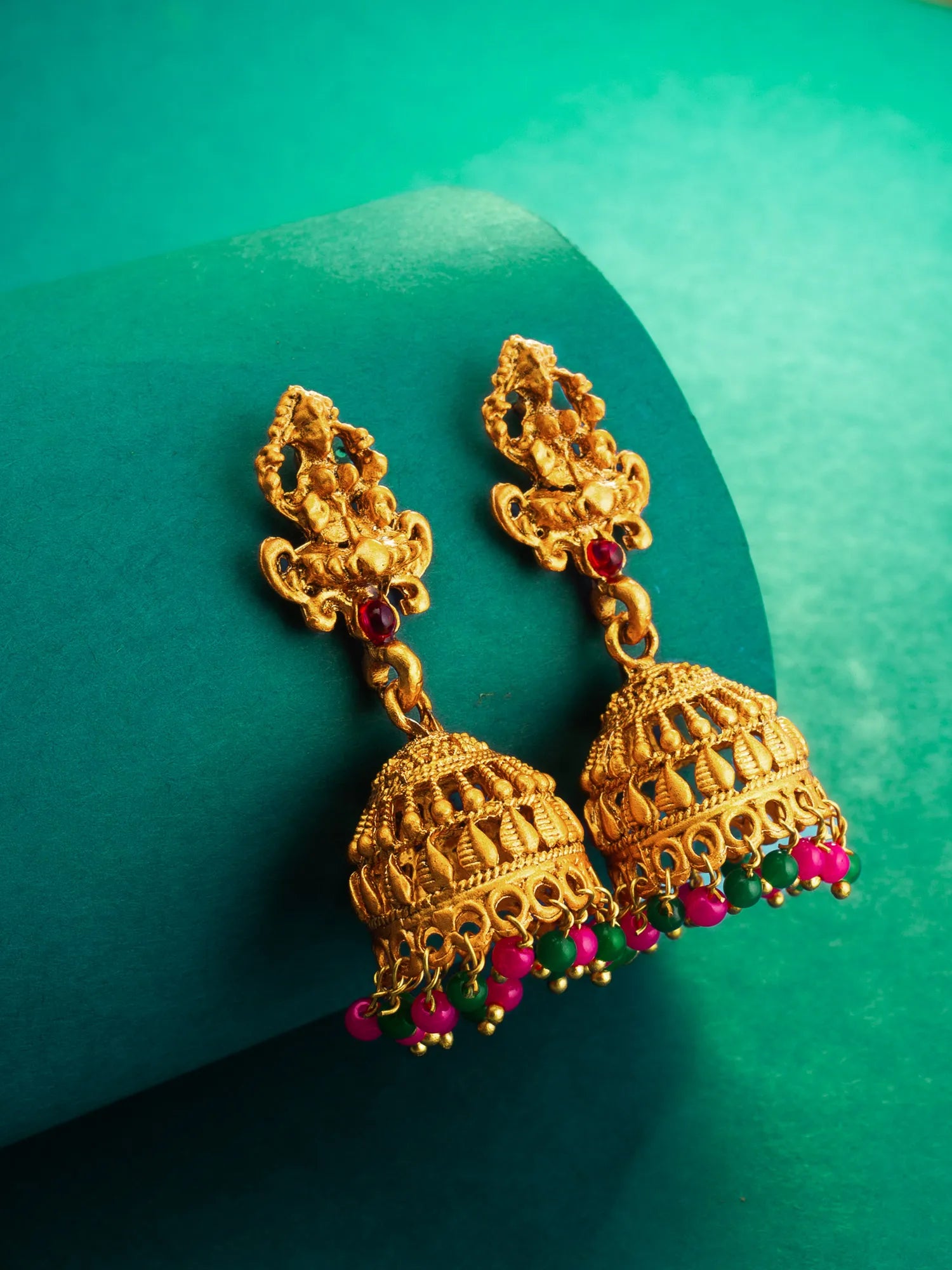 A pair of ornate gold-toned jhumka earrings with small red and green stone accents and dangling pink bead drops, displayed on a curved teal surface.