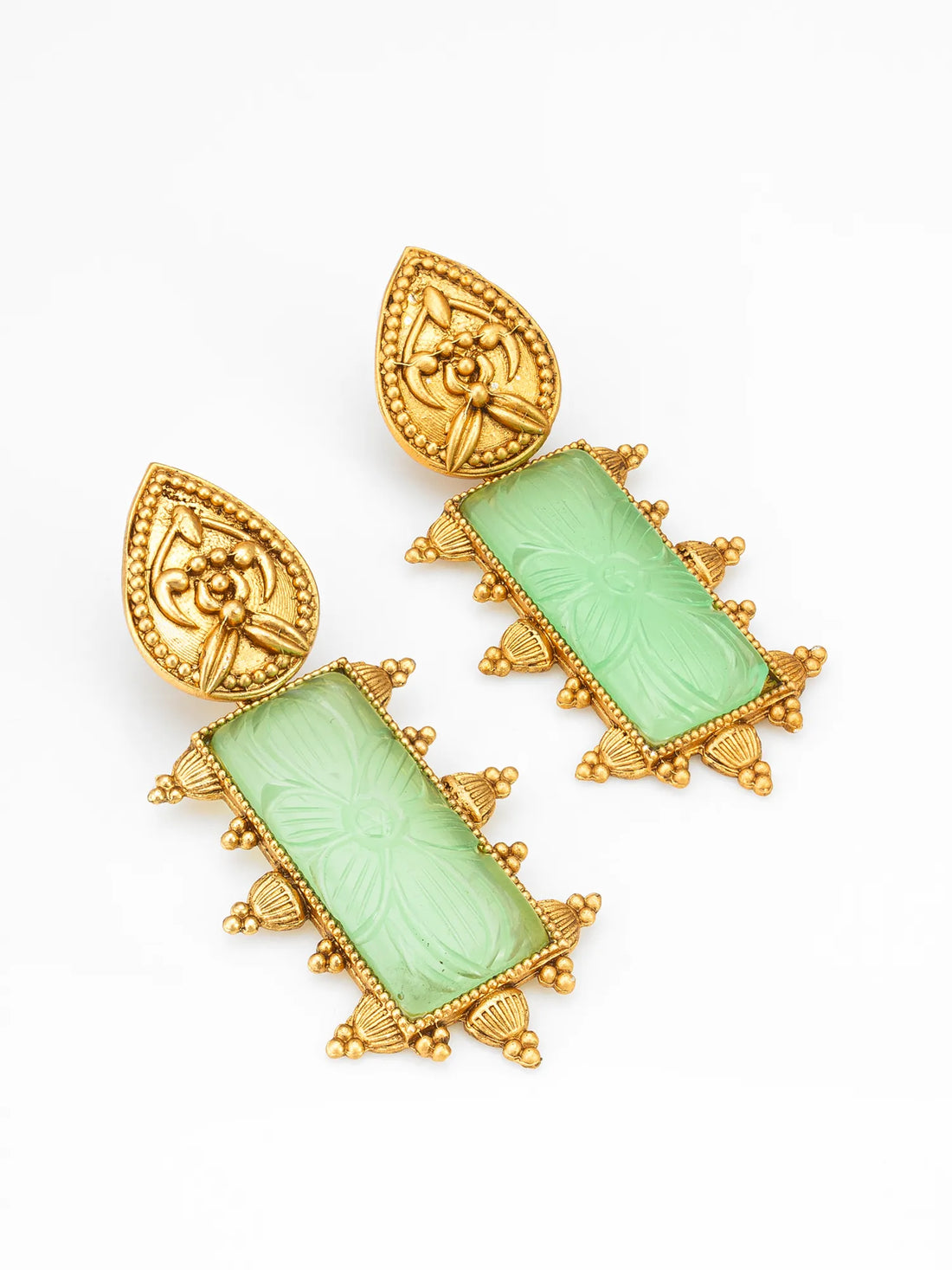 A pair of ornate gold-toned earrings with carved light green rectangular stone drops, displayed on a green surface with a blurred pink bowl in the background.