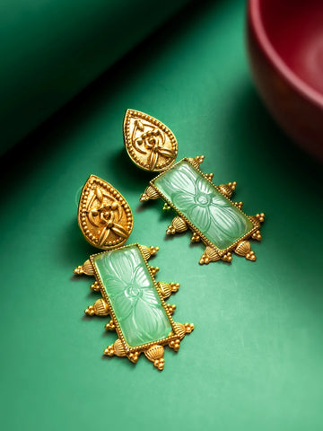 A pair of ornate gold-toned earrings with carved light green rectangular stone drops, displayed on a green surface with a blurred pink bowl in the background.
