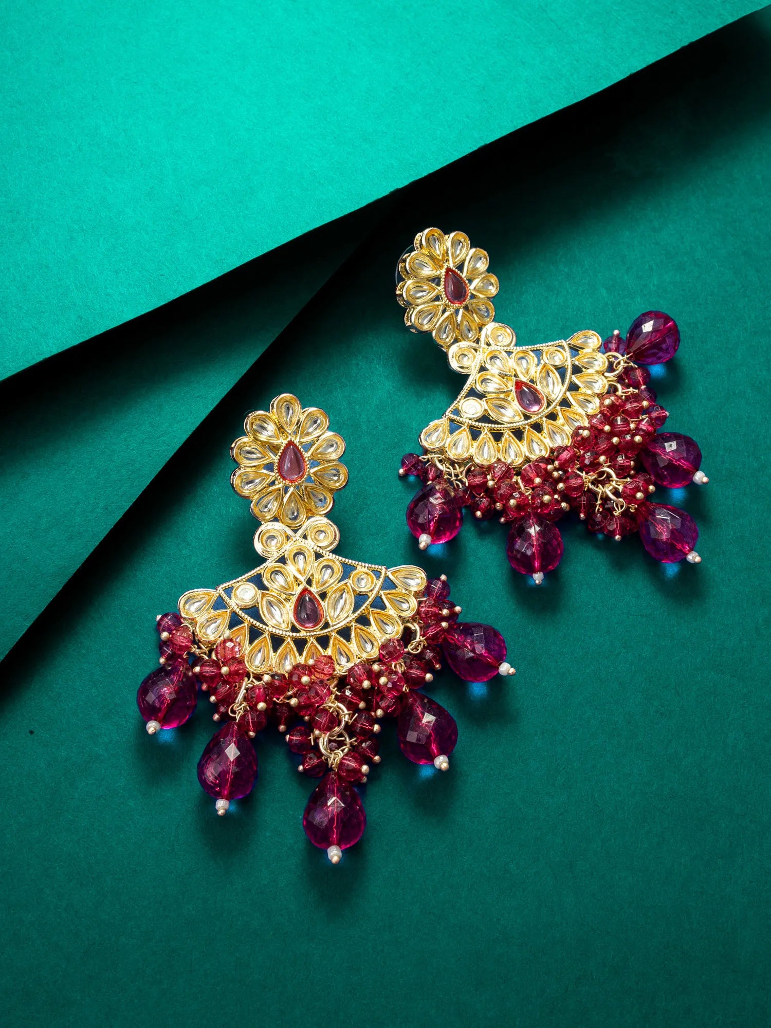A pair of ornate gold-plated Indian jhumka earrings decorated with deep red beads and purple gemstone accents, displayed on a dark green background.