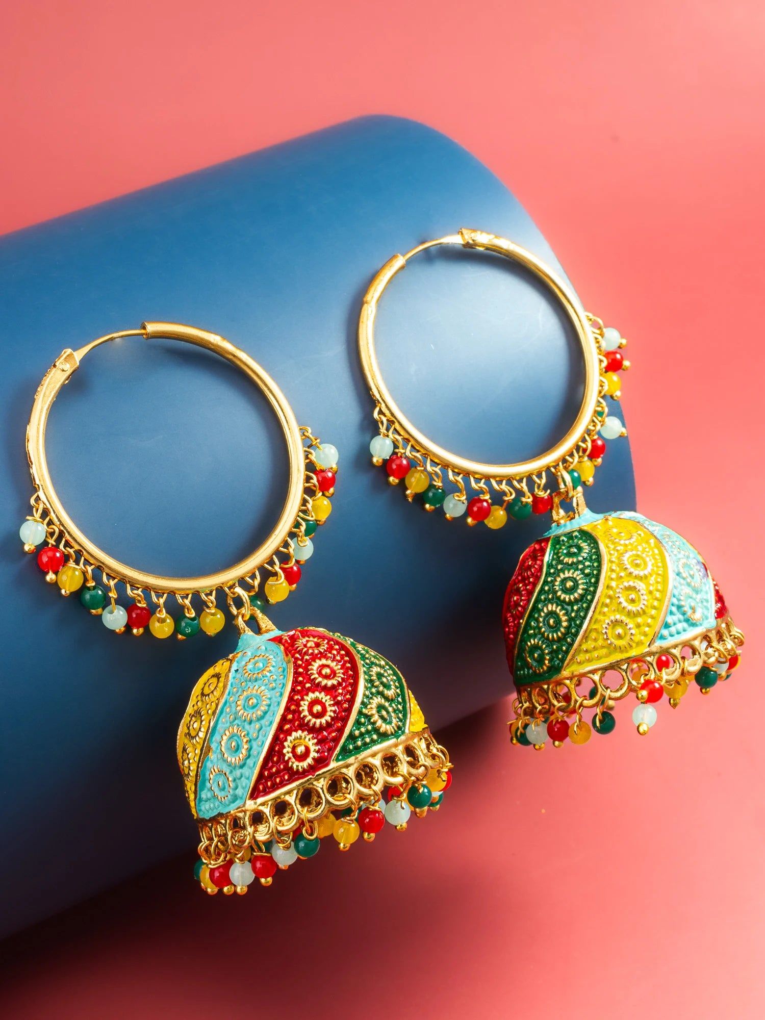 A pair of gold-tone hoop earrings with dangling multicolor jhumka-style charms, displayed on a blue cylindrical prop against a pink background.