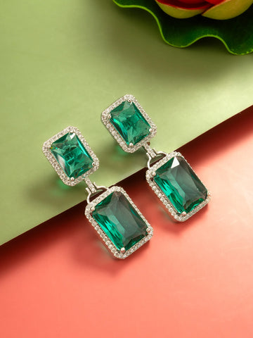 A pair of rectangular drop earrings with emerald-cut green stones surrounded by small clear stones in silver-toned settings, displayed on a split green and coral background.
