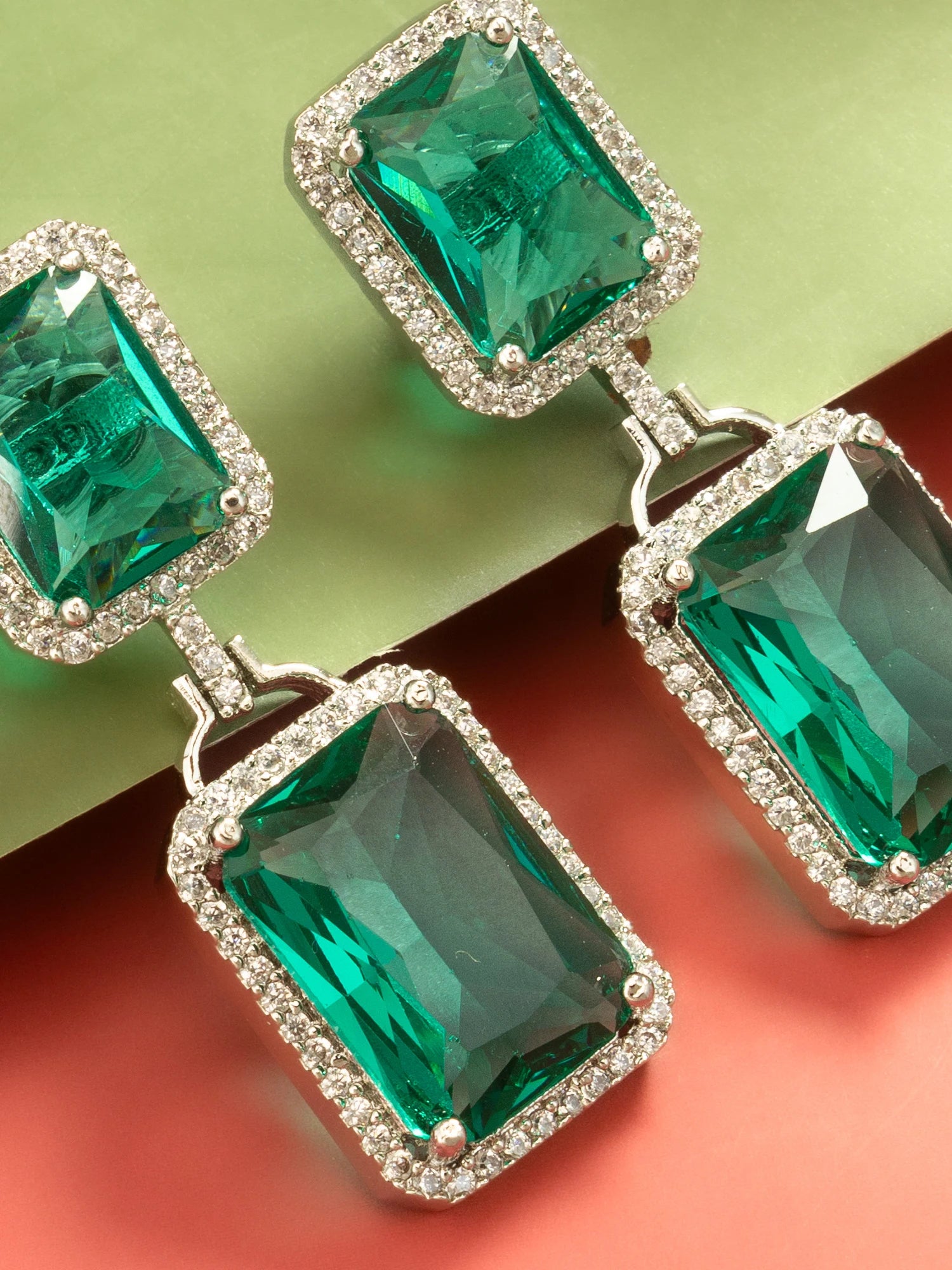 A pair of rectangular drop earrings featuring large emerald-cut green gemstones surrounded by small clear stones in a silver-tone setting, displayed on a split green and coral background.