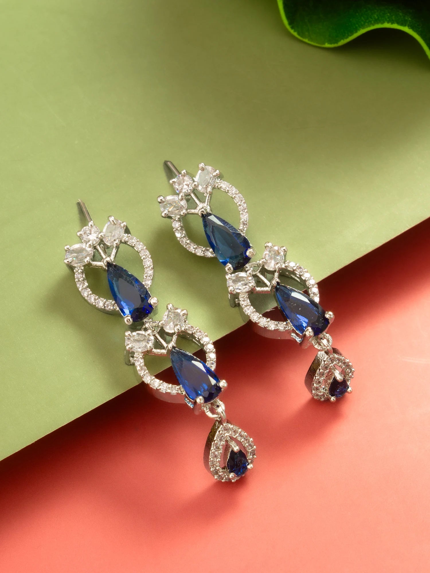 A pair of silver-tone drop earrings featuring deep blue teardrop-shaped gemstones and clear pavé-set accent stones, displayed on a split green and coral background.
