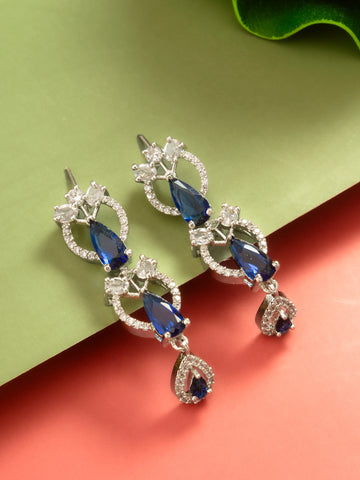 A pair of silver-tone drop earrings featuring deep blue teardrop-shaped gemstones and clear pavé-set accent stones, displayed on a split green and coral background.
