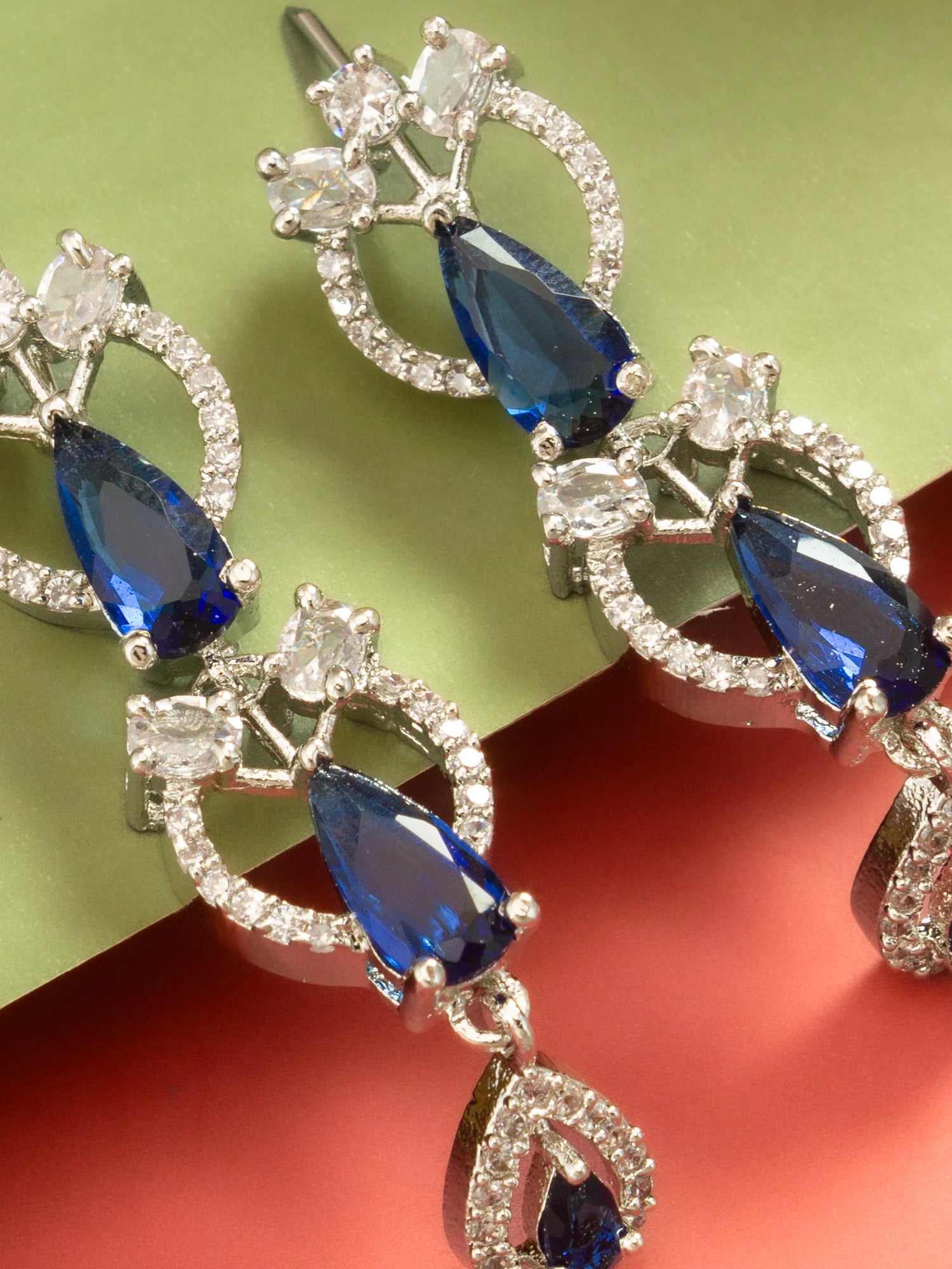 A pair of silver-tone drop earrings featuring teardrop-shaped blue gemstones and clear crystal accents, displayed on a split green and red background.