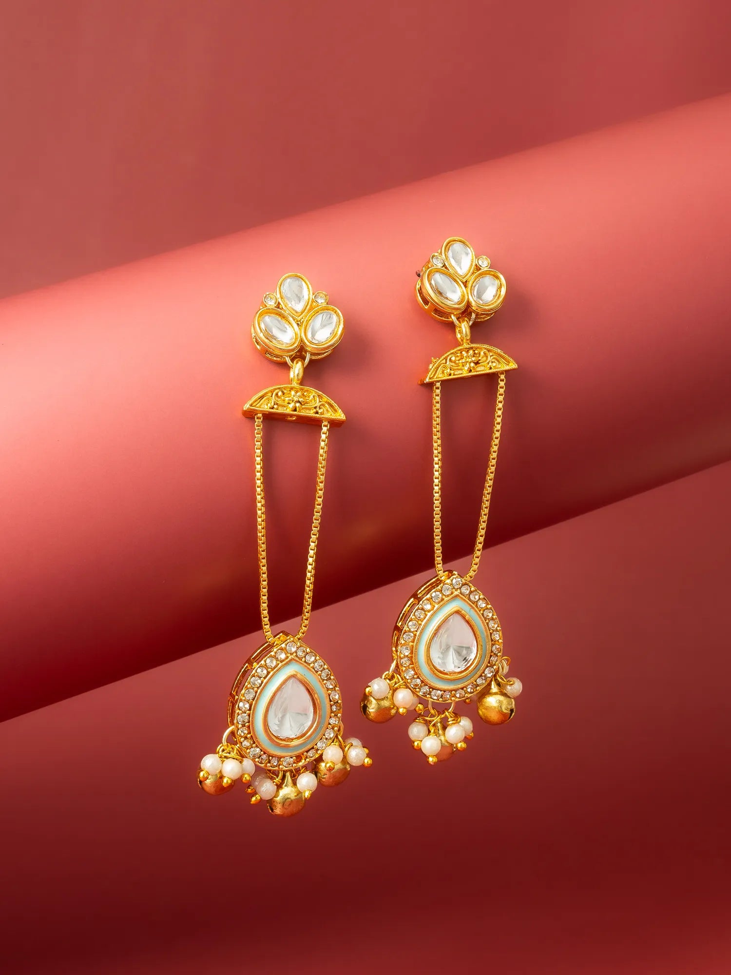 A pair of gold-toned Indian style dangle earrings with white faux pearls, displayed on a curved red background.