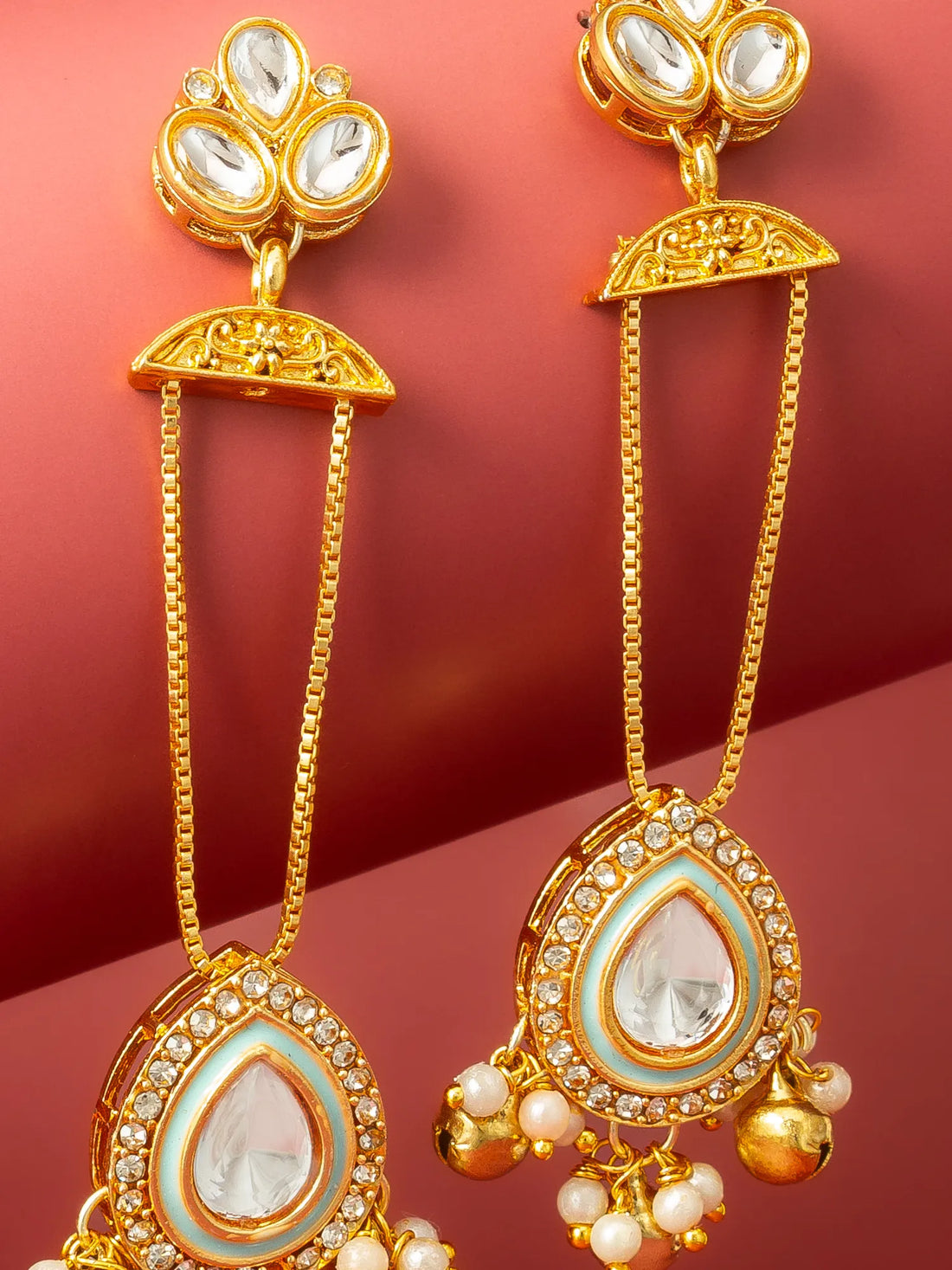 A pair of gold-toned Indian style dangle earrings with white faux pearls, displayed on a curved red background.