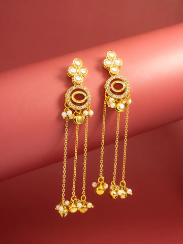 A pair of gold-toned traditional Indian dangle earrings with white pearl-like beads and clear rhinestone accents, displayed on a curved pinkish-red background.
