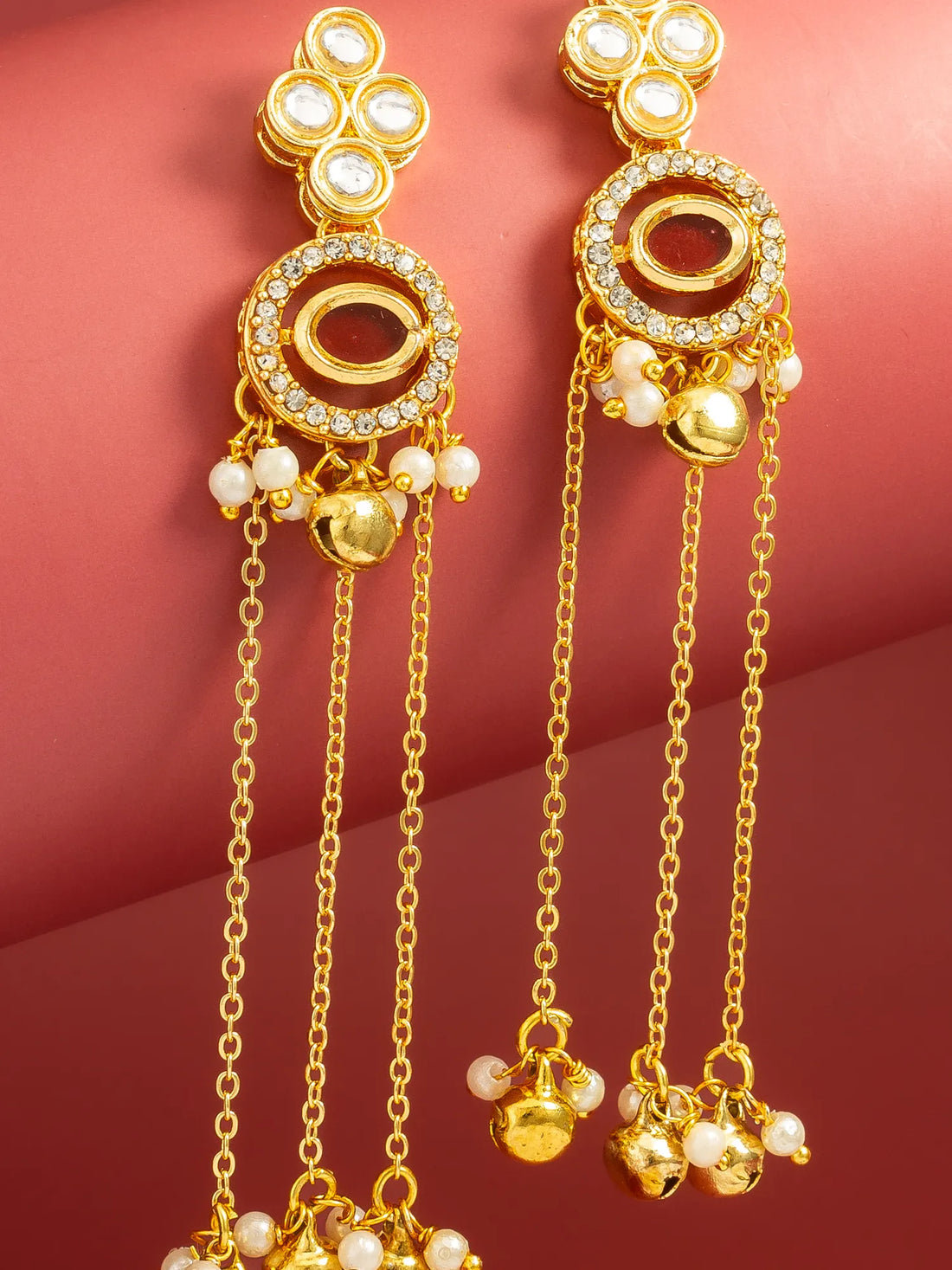 A pair of gold-toned traditional Indian dangle earrings with white pearl-like beads and clear rhinestone accents, displayed on a curved pinkish-red background.