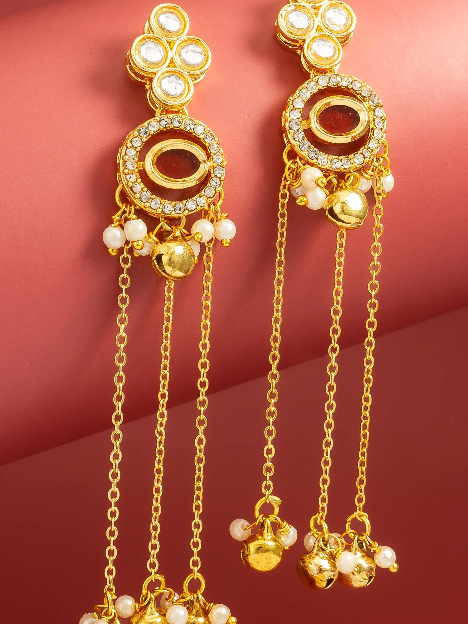 A pair of gold-toned traditional Indian dangle earrings with white pearl-like beads, clear rhinestones, and small gold bells, displayed on a warm pinkish-red background.