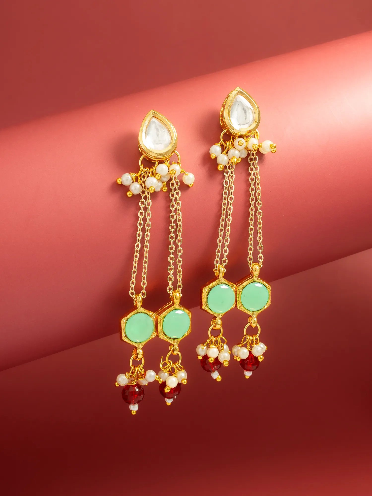 A pair of gold-plated Indian Kundan dangle earrings with white stones, green hexagonal stones, and red bead drops, displayed on a curved red background.