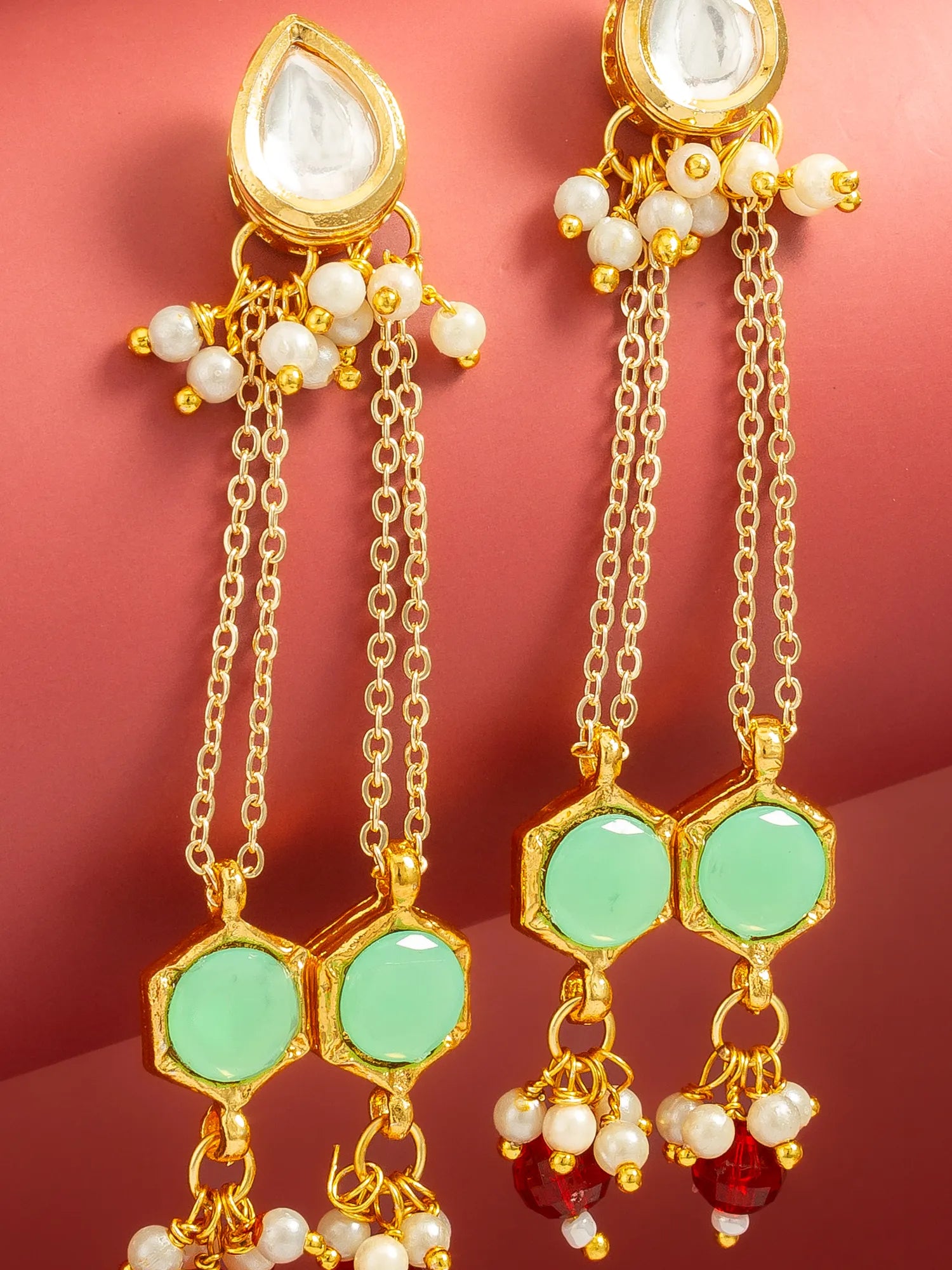 A pair of gold-toned traditional Indian dangle earrings with light green stone inlays, white pearl beads, and a clear teardrop stone at the top, displayed against a solid pink background.