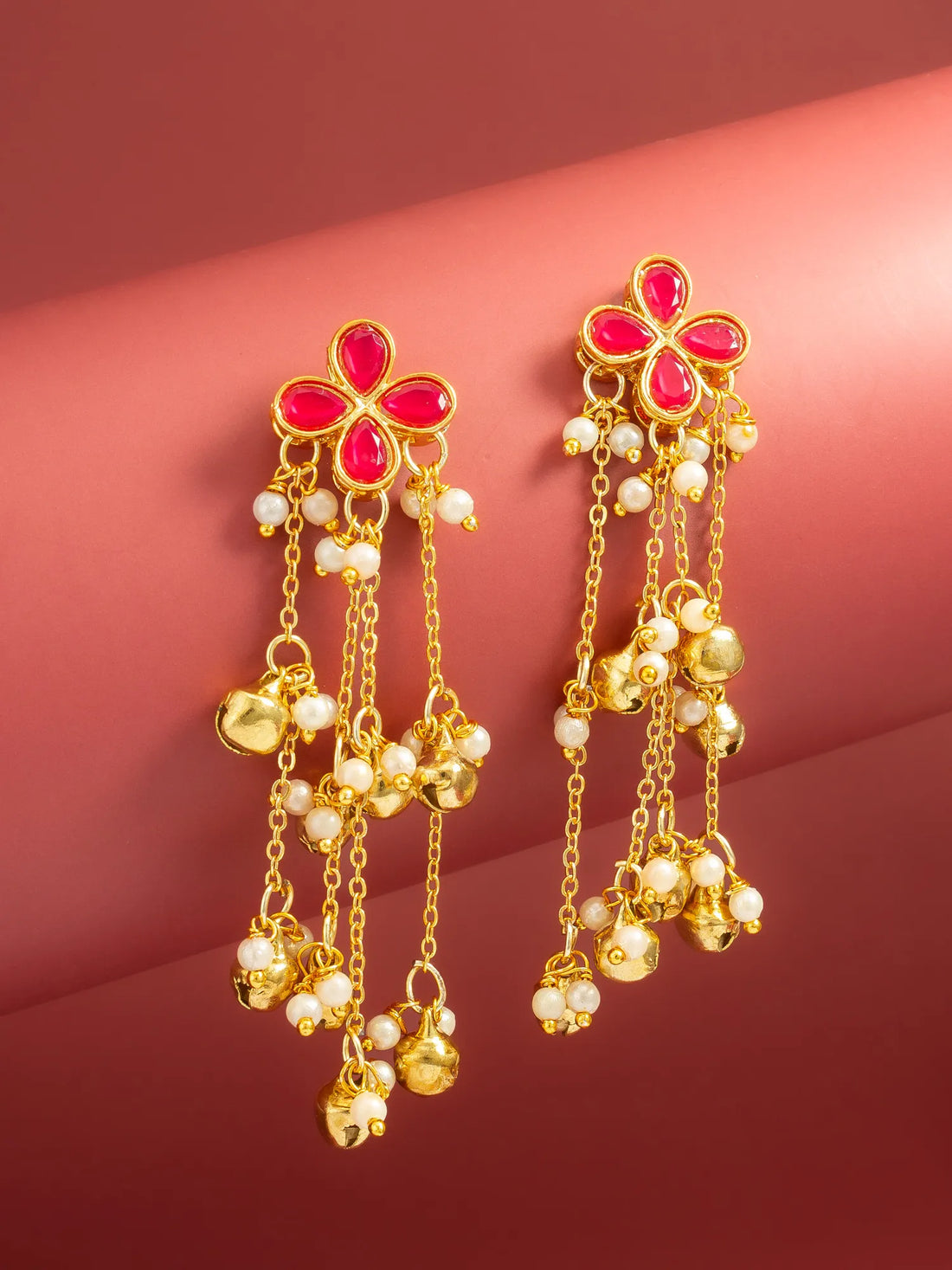 A pair of gold-toned traditional Indian earrings with red flower-shaped stones, white pearl beads, and dangling gold bells, displayed on a curved pinkish-red background.