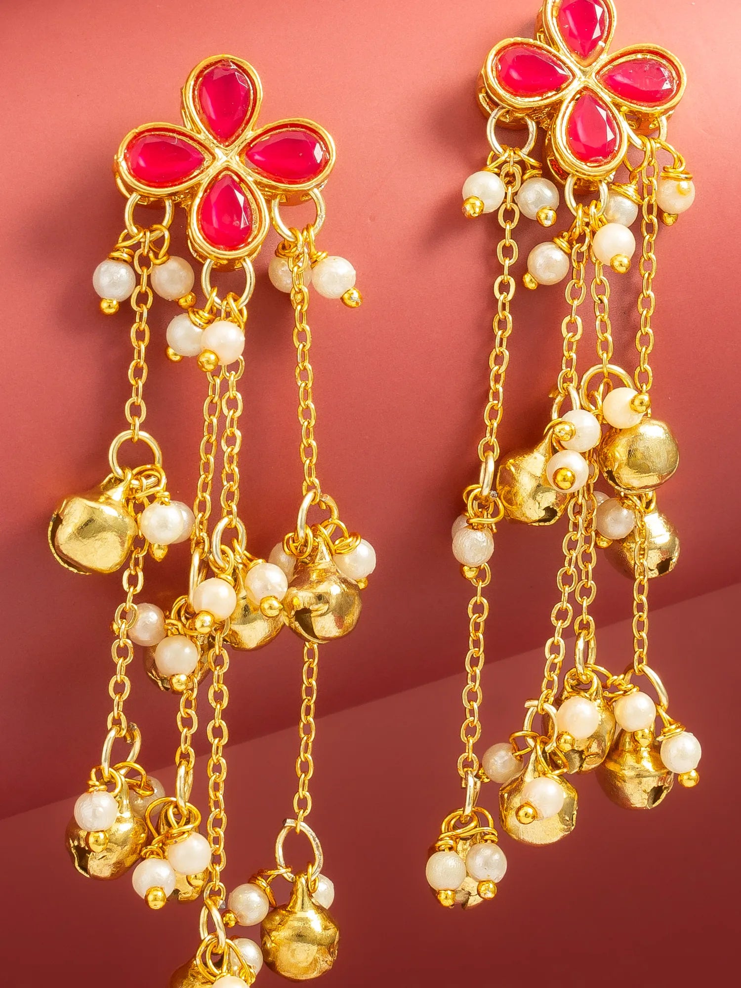 A pair of gold-toned traditional Indian dangle earrings with bright pink stone flower tops, white pearl beads, and small gold bells, shown against a solid pink background.