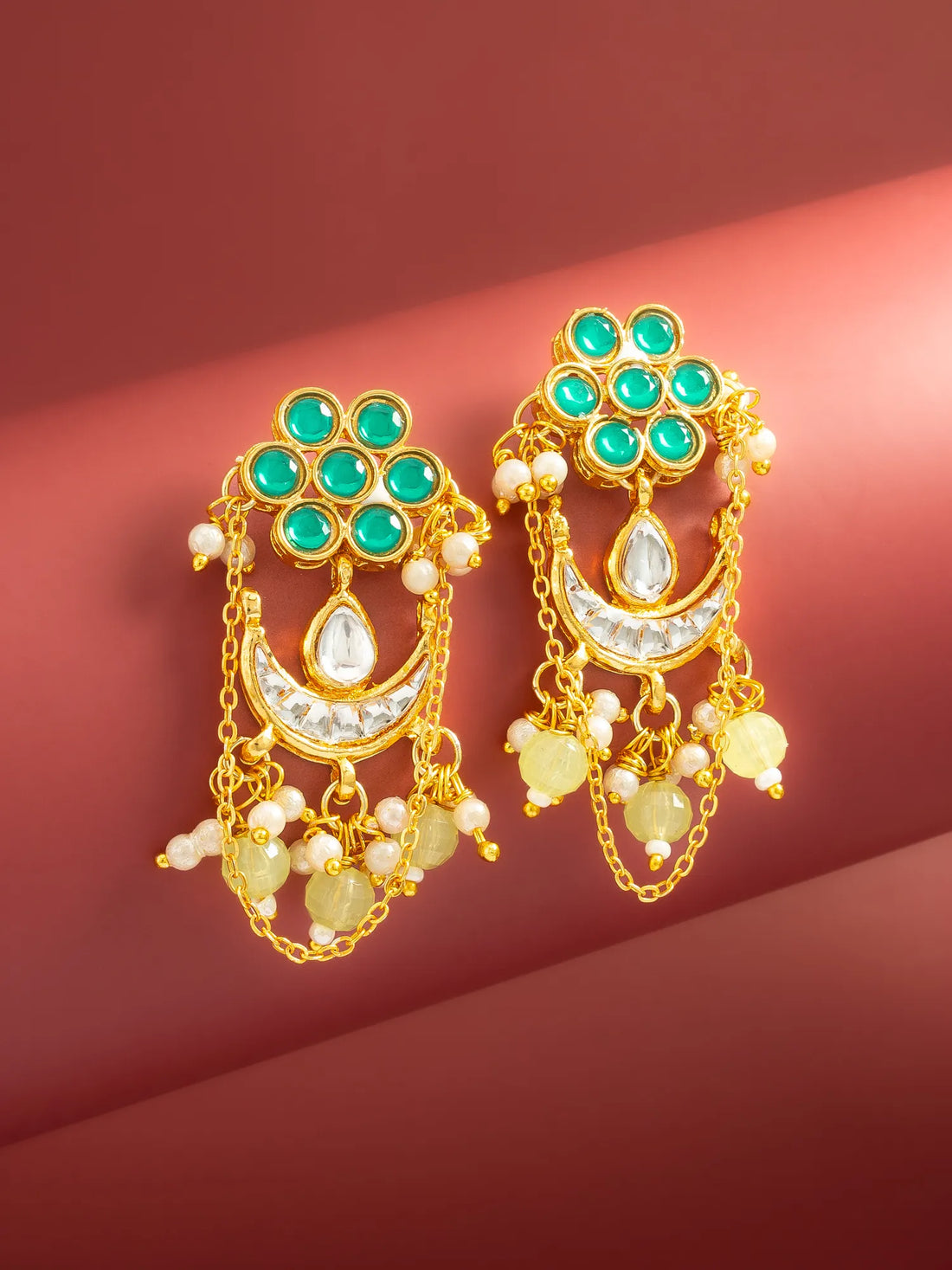 A pair of gold-toned traditional Indian earrings with green stones, white pearl-like beads, and yellow stone accents, displayed on a dark red background.