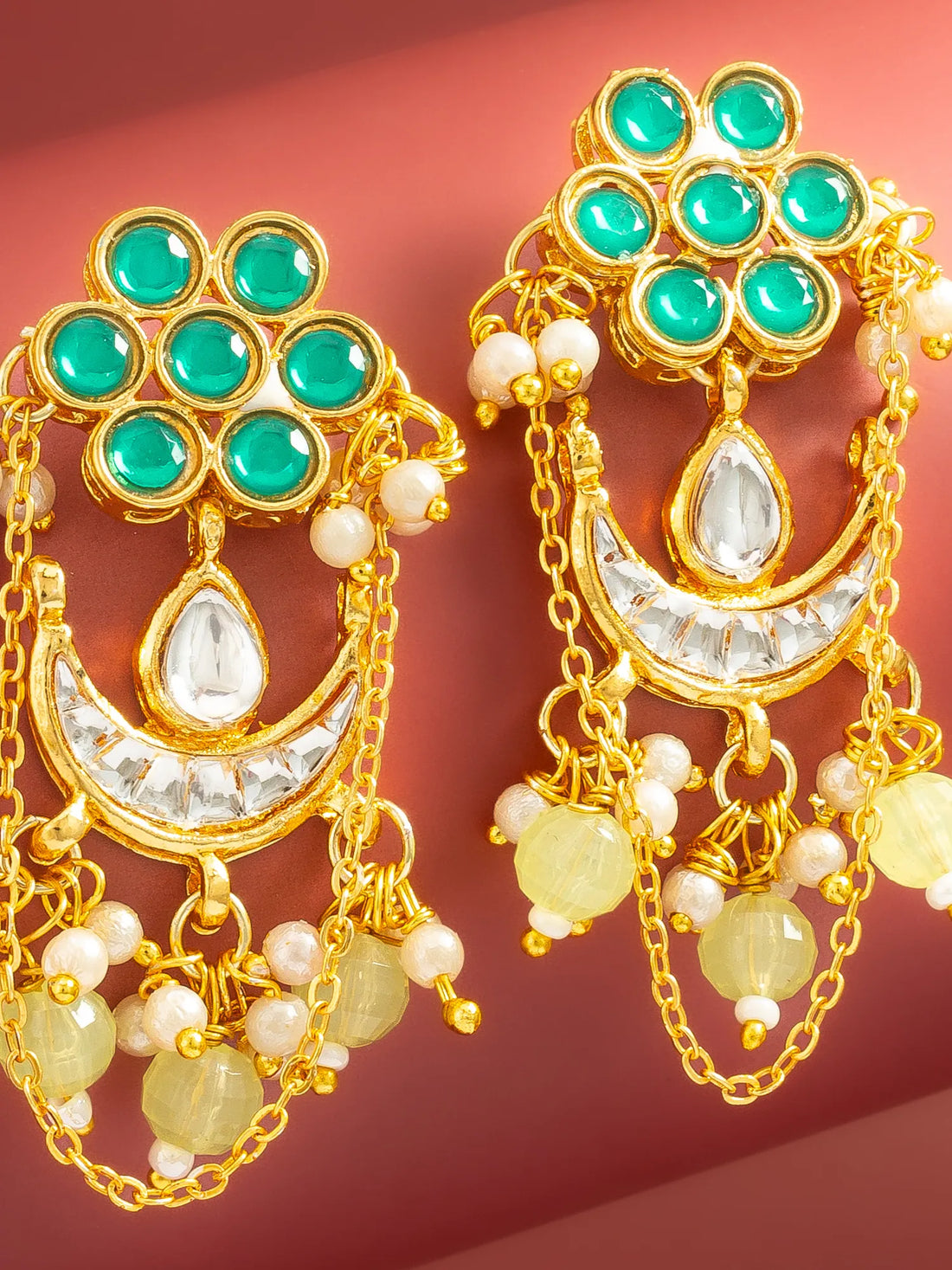 A pair of gold-toned traditional Indian earrings with green stones, white pearl-like beads, and yellow stone accents, displayed on a dark red background.