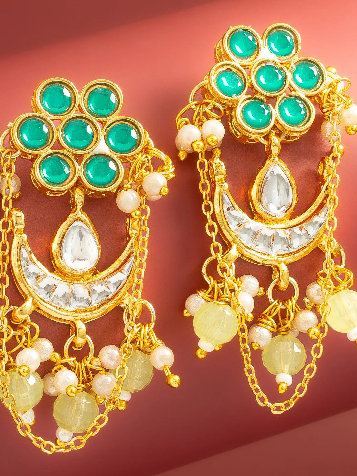A pair of ornate gold-toned Indian jhumka earrings featuring green stone clusters, a crescent moon with a clear teardrop stone, white pearl beads, and yellow-green bead drops, set against a solid reddish-brown background.