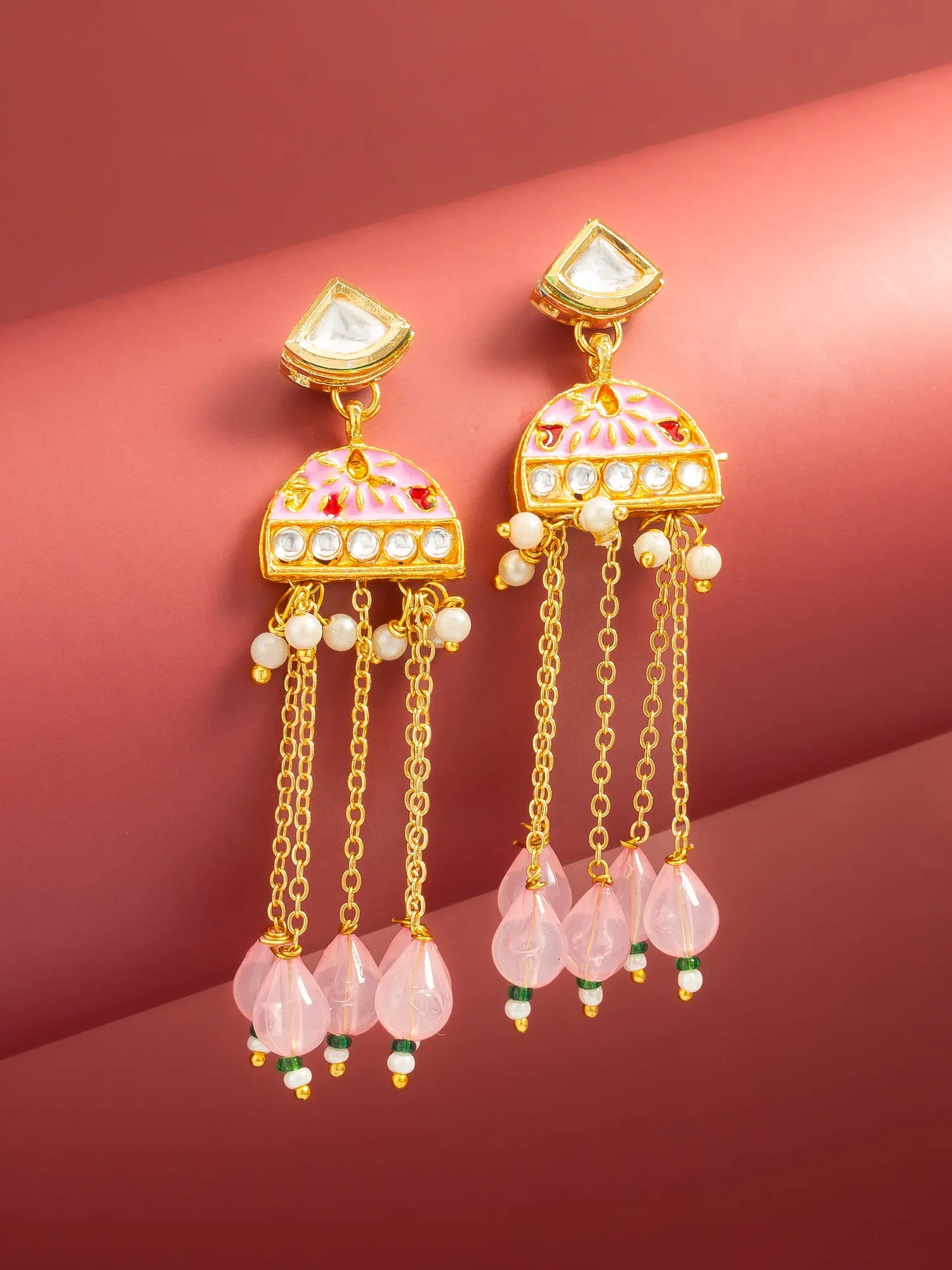 A pair of gold-toned traditional Indian jhumka earrings with pink enamel, white stones, and dangling pink teardrop beads, displayed on a curved pink background.