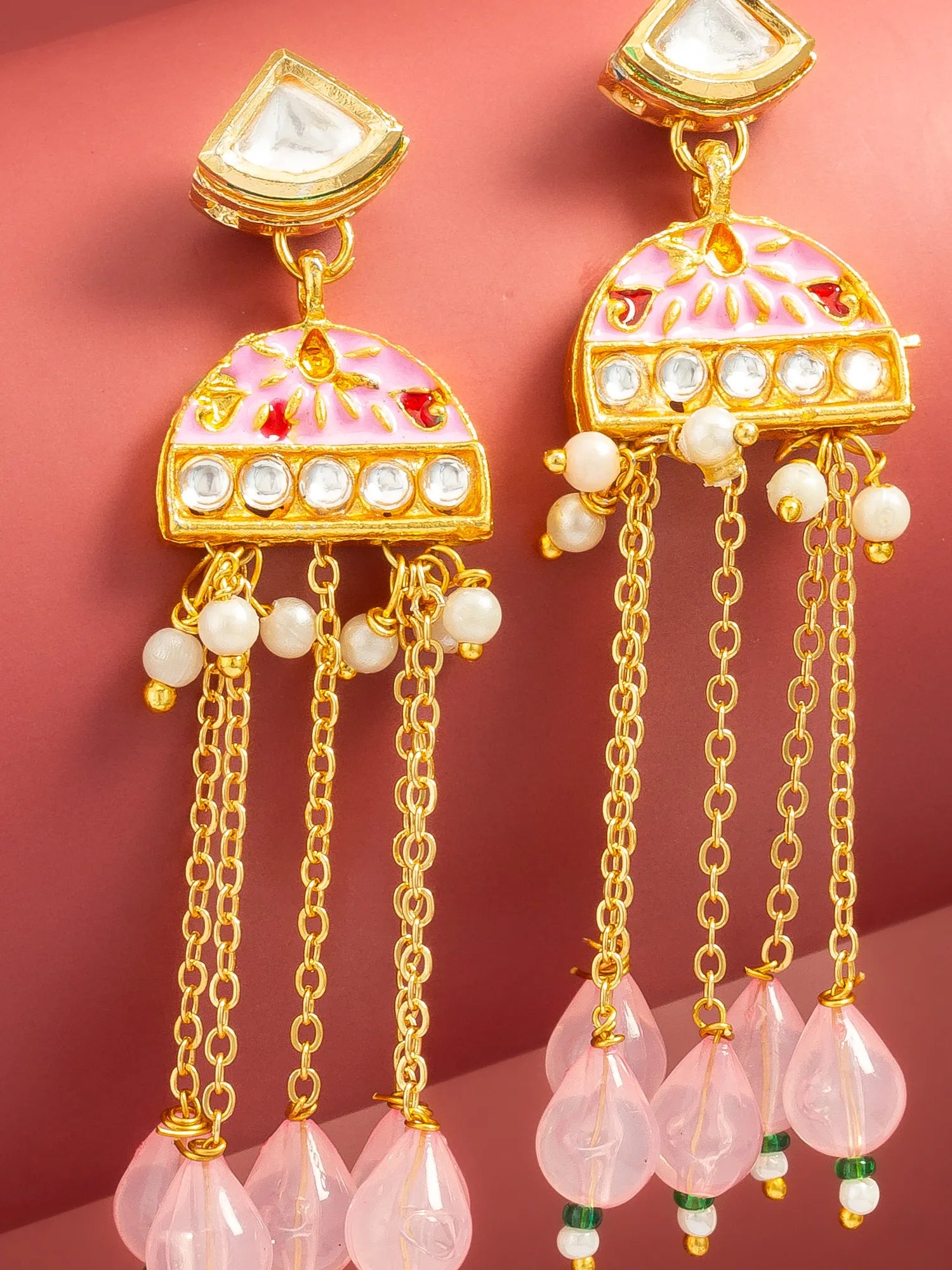 A pair of gold-plated Indian jhumka earrings with pink and white enamel detailing, white stones, and dangling pink teardrop beads, set against a solid pink background.
