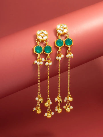 A pair of gold-toned dangle earrings with green stones and small white pearl-like accents, displayed against a warm pinkish-red studio background.