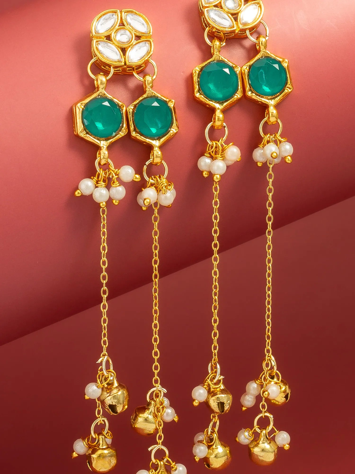 Gold-toned traditional Indian dangle earrings with green stones, white pearl beads, and hanging bell charms, displayed against a warm pink background.