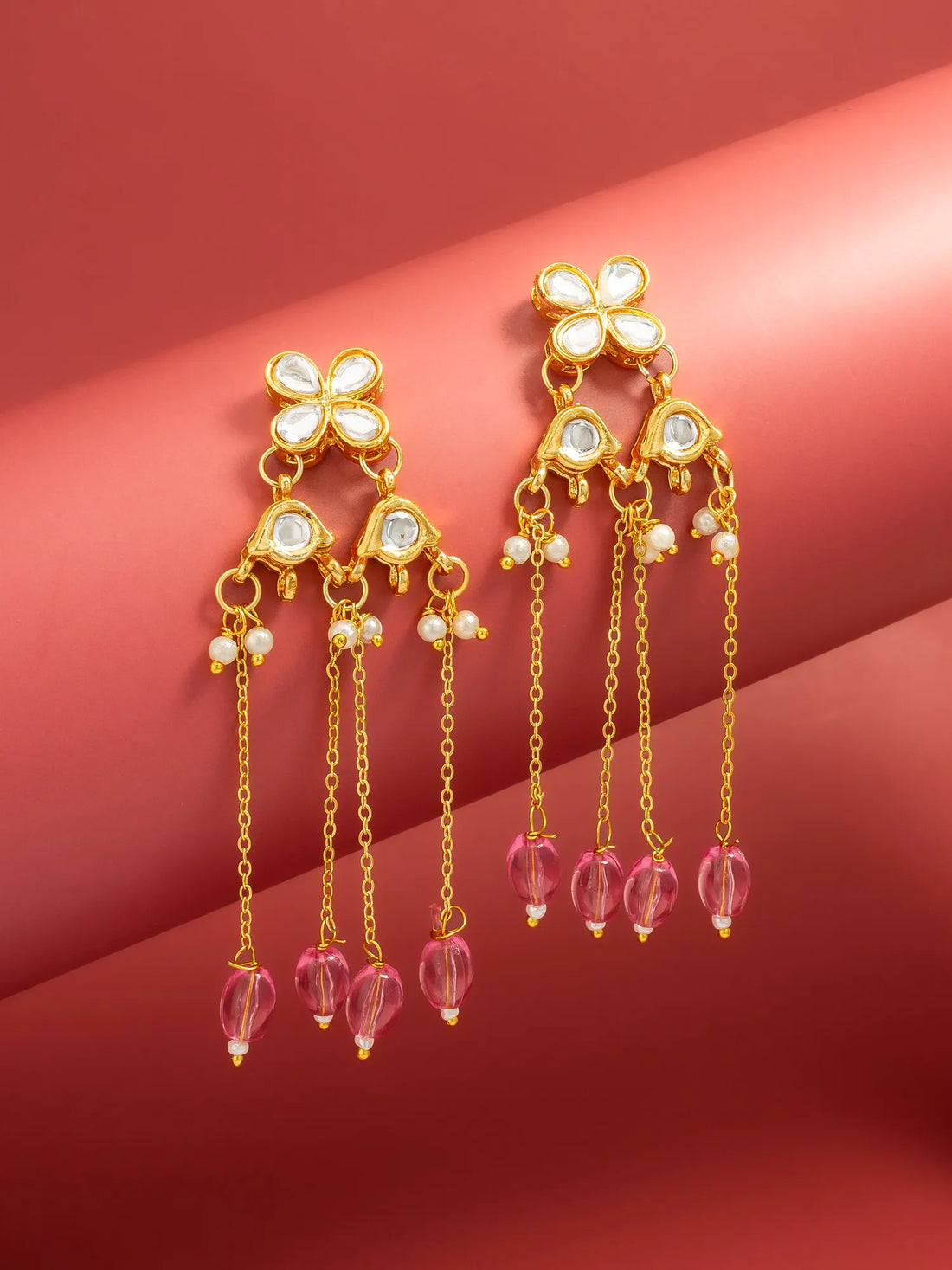 A pair of gold-toned dangle earrings with white stone floral tops, small pearl-like beads, and dangling pink faceted beads, displayed on a warm pink background.