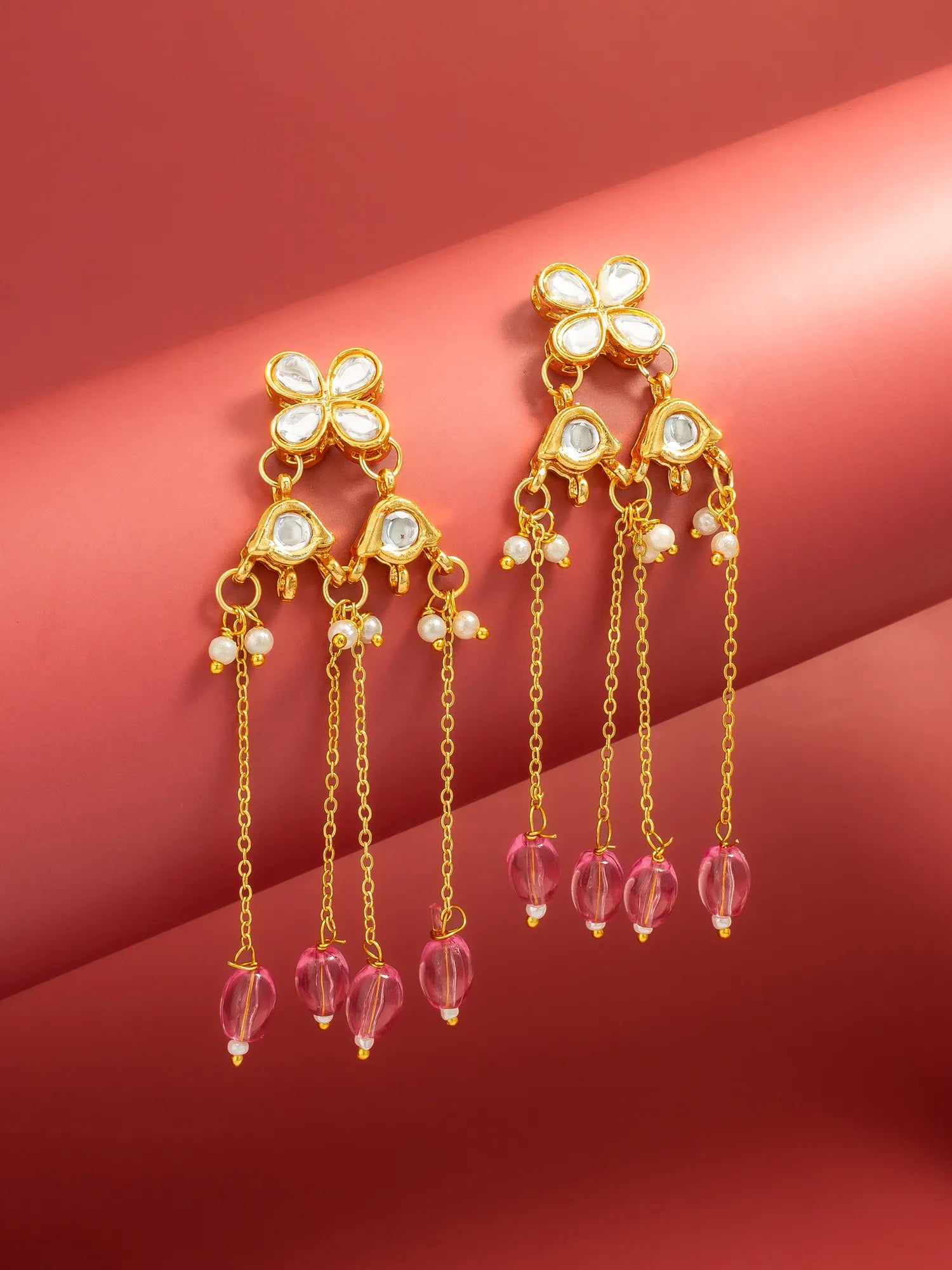 A pair of gold-toned dangle earrings with white stone floral tops, small pearl-like beads, and dangling pink faceted beads, displayed on a warm pink background.