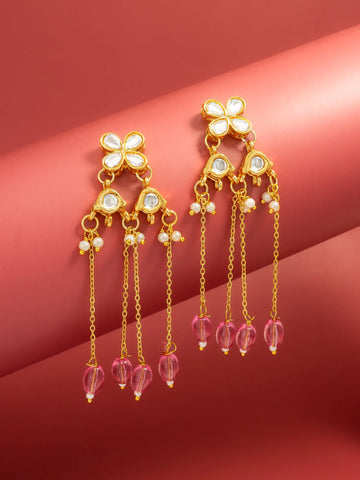 A pair of gold-toned dangle earrings with white stone floral tops, small pearl-like beads, and dangling pink faceted beads, displayed on a warm pink background.