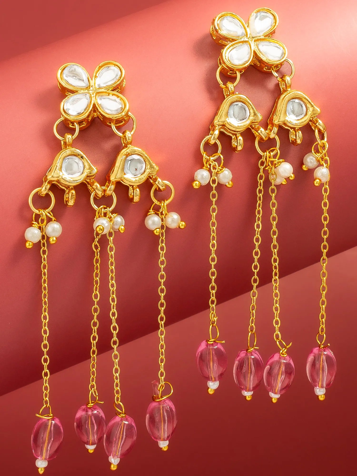 A pair of gold-toned chandelier earrings with white inlay stones, small pearl-like beads, and dangling pink faceted beads on a warm red background.
