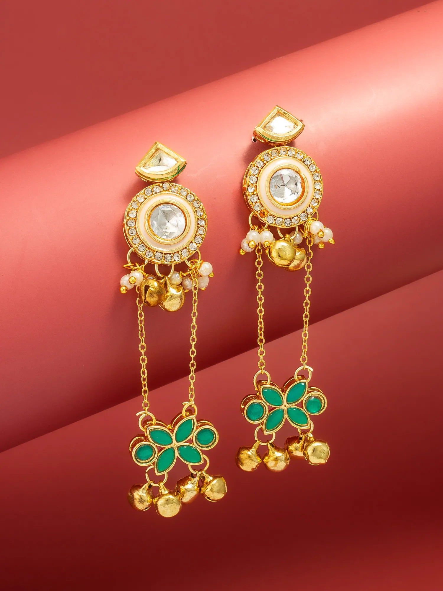 A pair of gold-toned Indian-style dangle earrings with green stone floral motifs, clear central stones, and small gold bells, displayed on a curved red background.