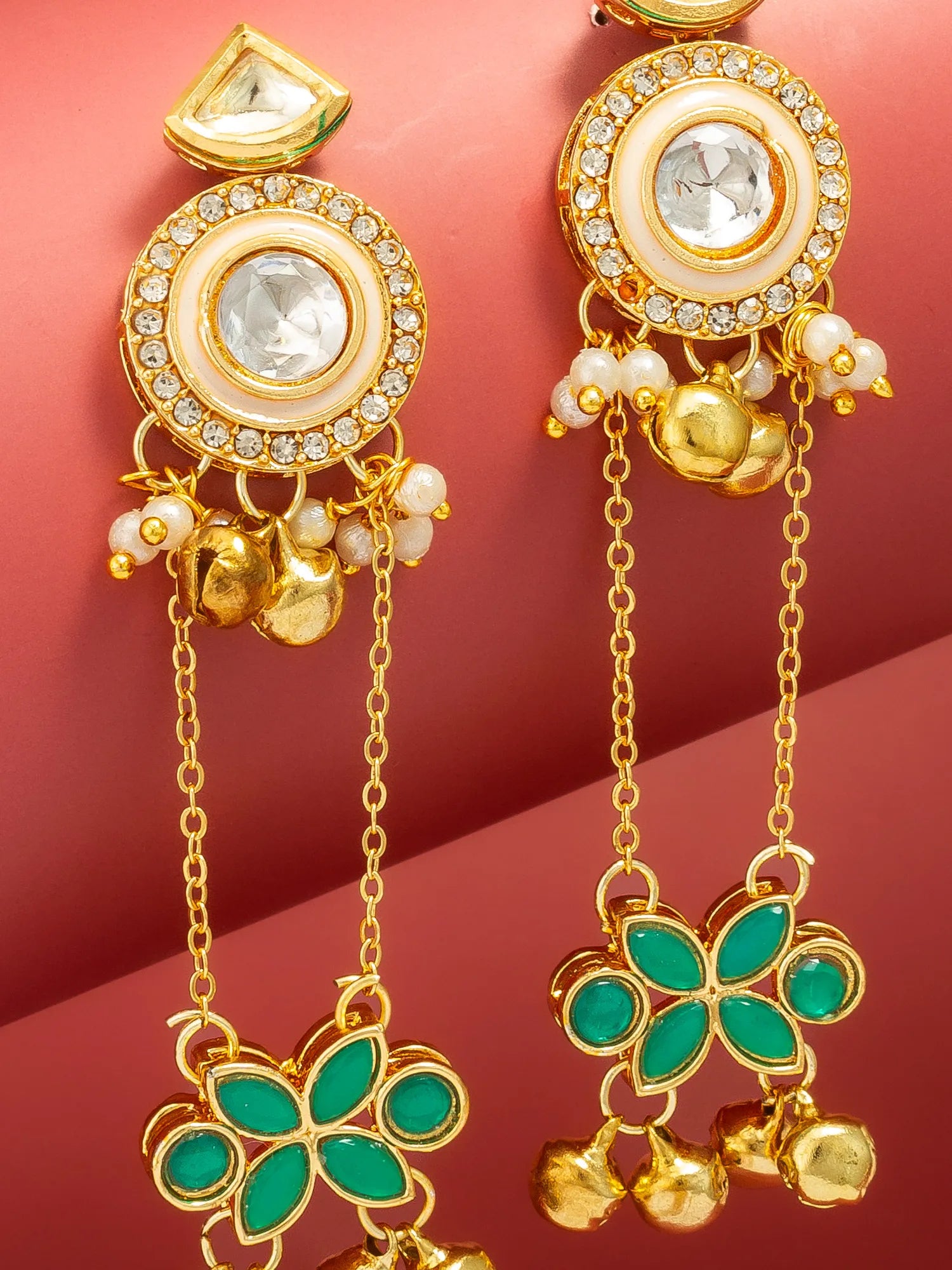 Gold-toned traditional Indian dangle earrings with clear stones, white pearl-like beads, and green enamel flower motifs, displayed on a pink background.