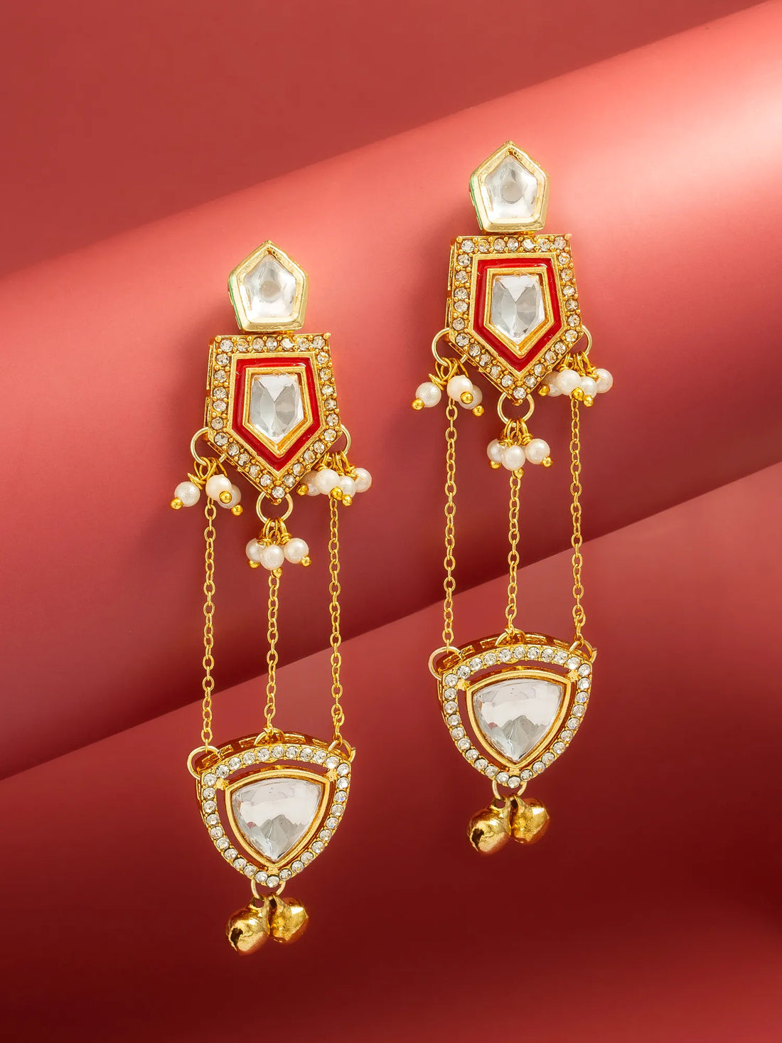 A pair of gold-toned traditional Indian dangle earrings with white pearl-like beads and clear rhinestone accents, displayed on a curved pinkish-red background.