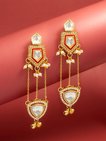 A pair of gold-toned traditional Indian dangle earrings with white pearl-like beads and clear rhinestone accents, displayed on a curved pinkish-red background.