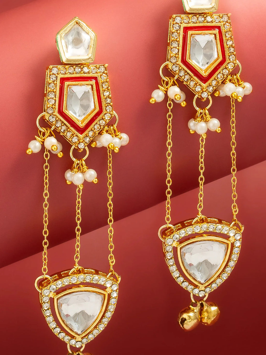 A pair of gold-toned traditional Indian dangle earrings with white pearl-like beads and clear rhinestone accents, displayed on a curved pinkish-red background.