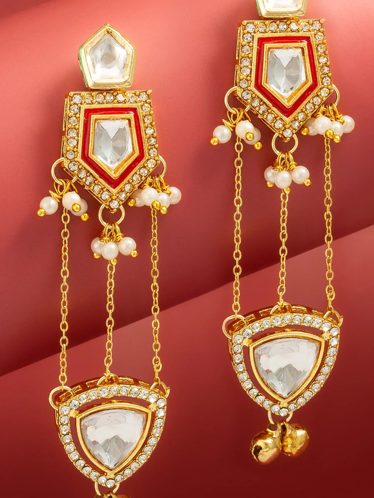 A pair of gold-toned traditional Indian dangle earrings with white pearl-like beads, clear rhinestones, and small gold bells, displayed on a warm pinkish-red background.