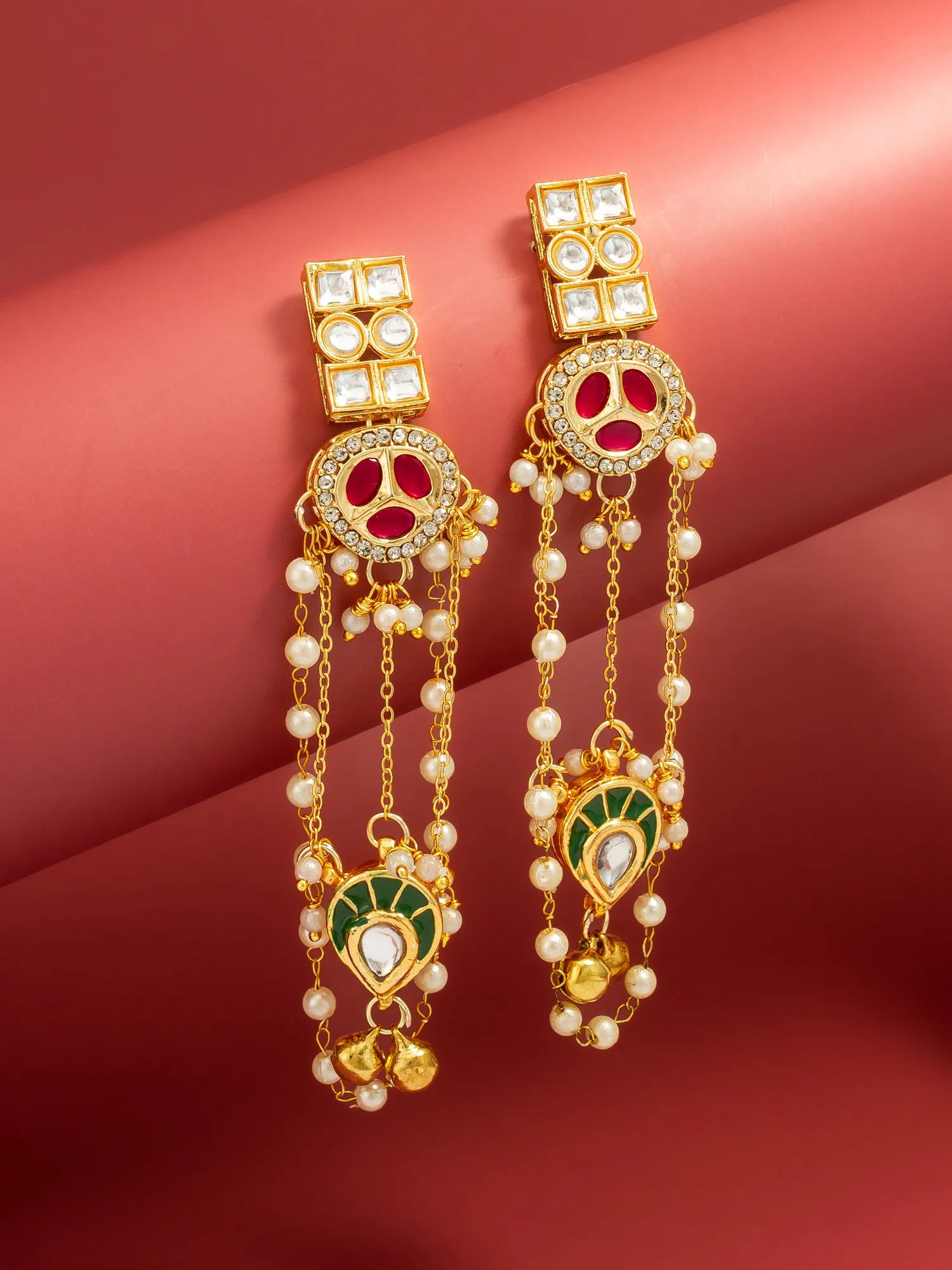A pair of gold-toned traditional Indian chandelier earrings decorated with white faux pearls, red enamel, green enamel, and clear stones, displayed on a pink background.