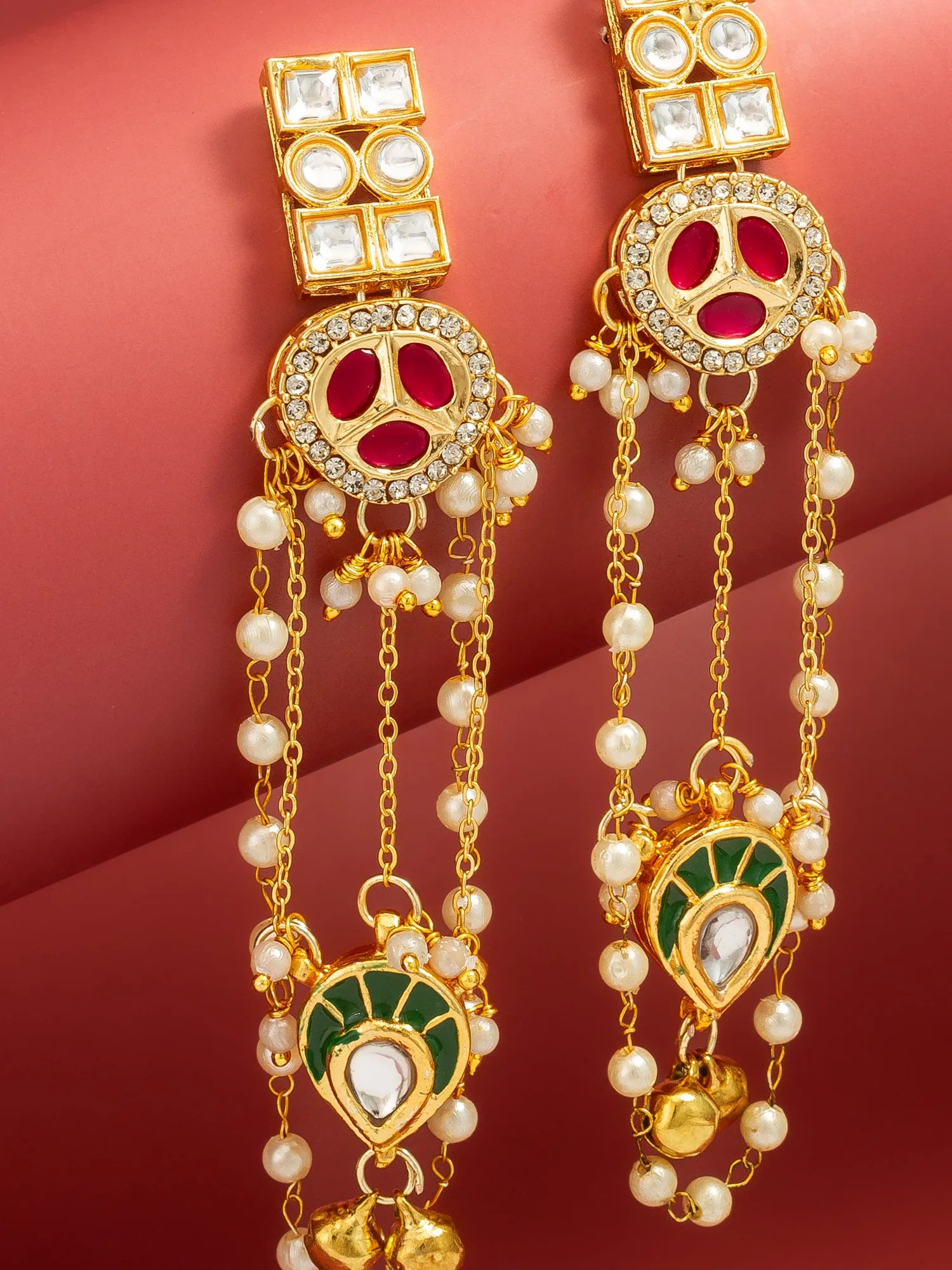 A pair of gold-plated Indian Kundan dangle earrings with red enamel, green enamel, white faux pearls, and clear crystals, displayed against a dark red background.