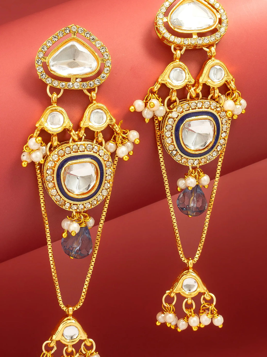 A pair of ornate gold-plated dangle earrings with white stones, small pearl-like bead accents, and purple teardrop charms, displayed against a red background.