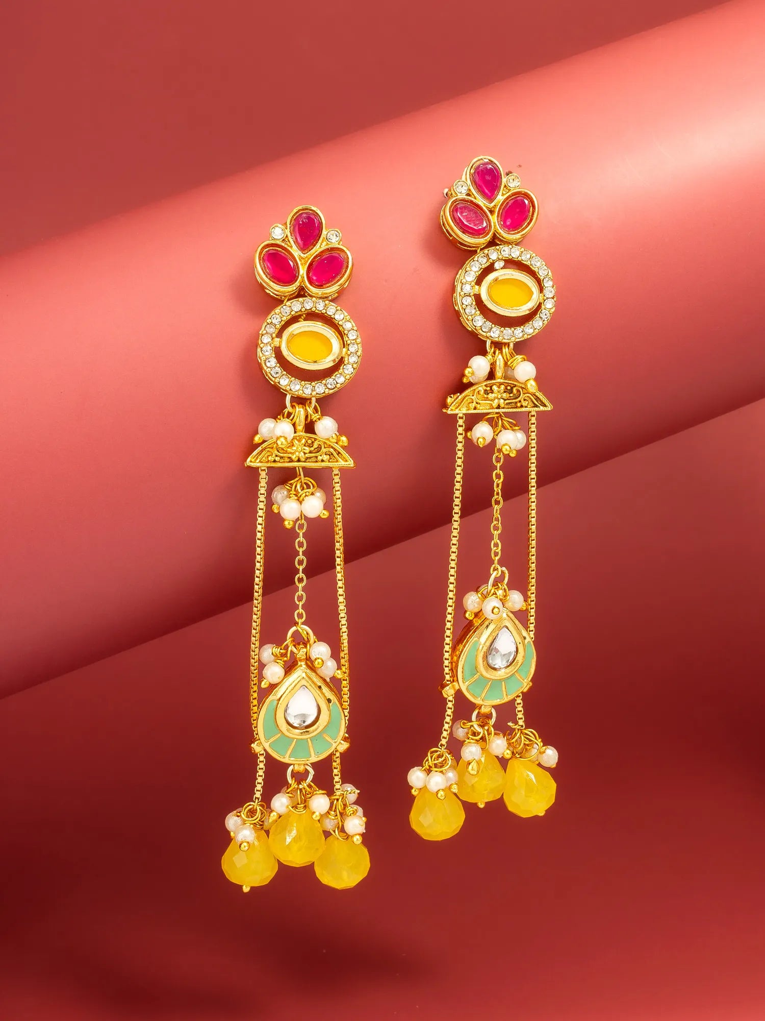 A pair of gold-toned Indian jhumka earrings with red, yellow, and green stones, set against a pinkish-red background.